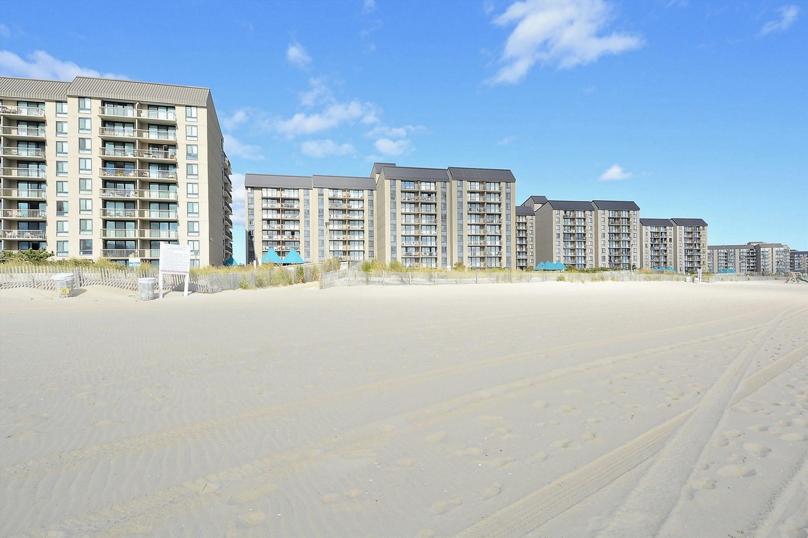 Modern beachfront buildings line pristine sandy shores under clear blue skies with gentle sea grass dunes.