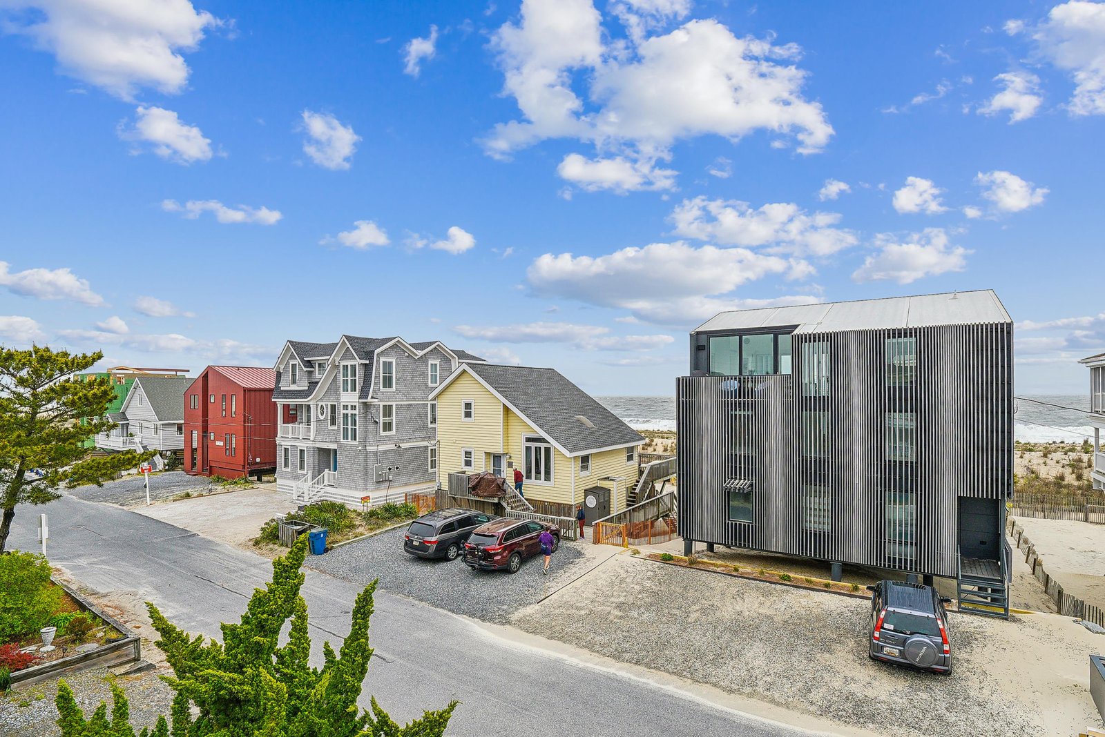 A modern coastal property with distinctive metal siding, surrounded by neighboring beach houses and situated near the sandy shoreline.