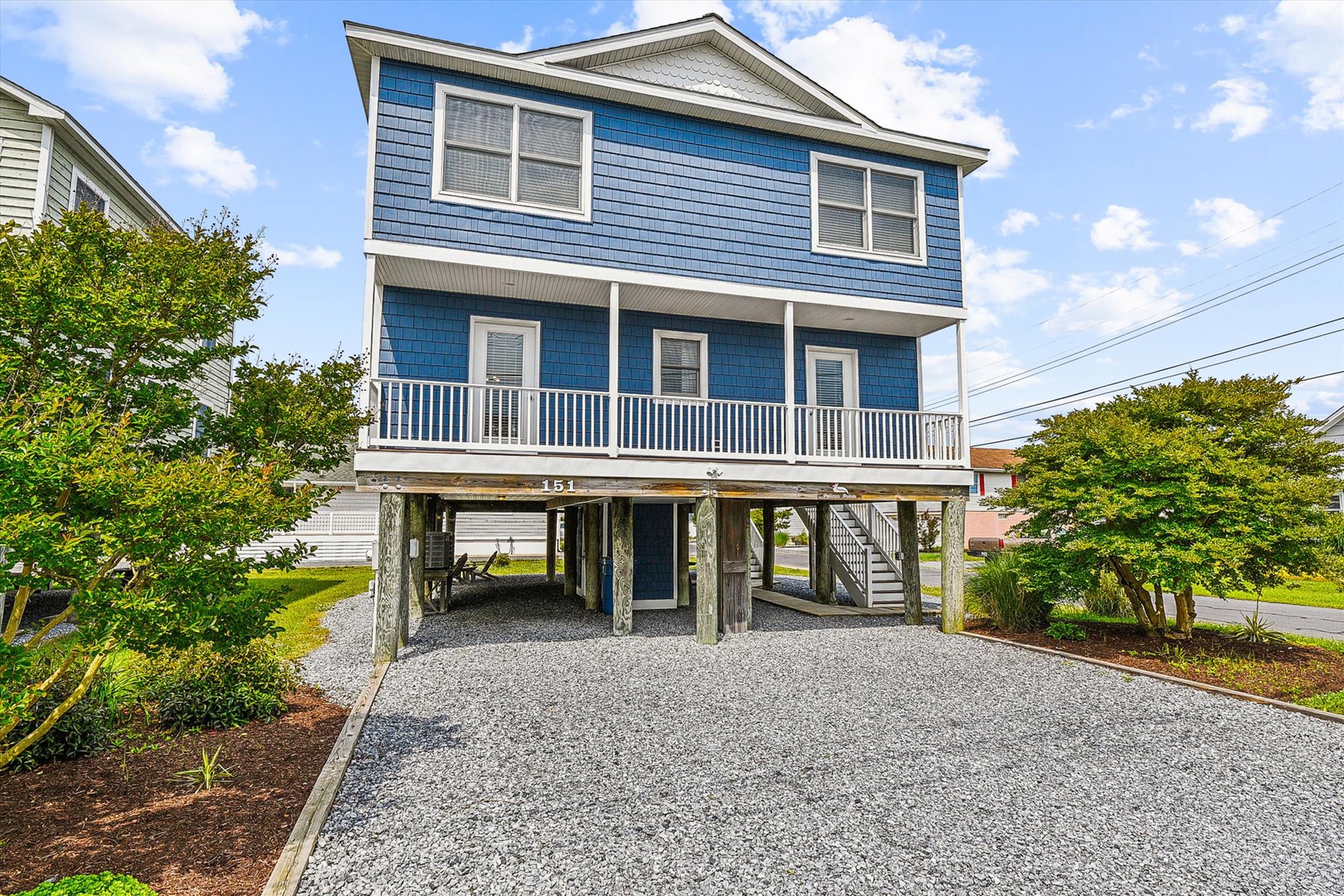 Coastal-style two-story home elevated on stilts with a spacious front porch, surrounded by lush greenery and offering convenient gravel parking below.