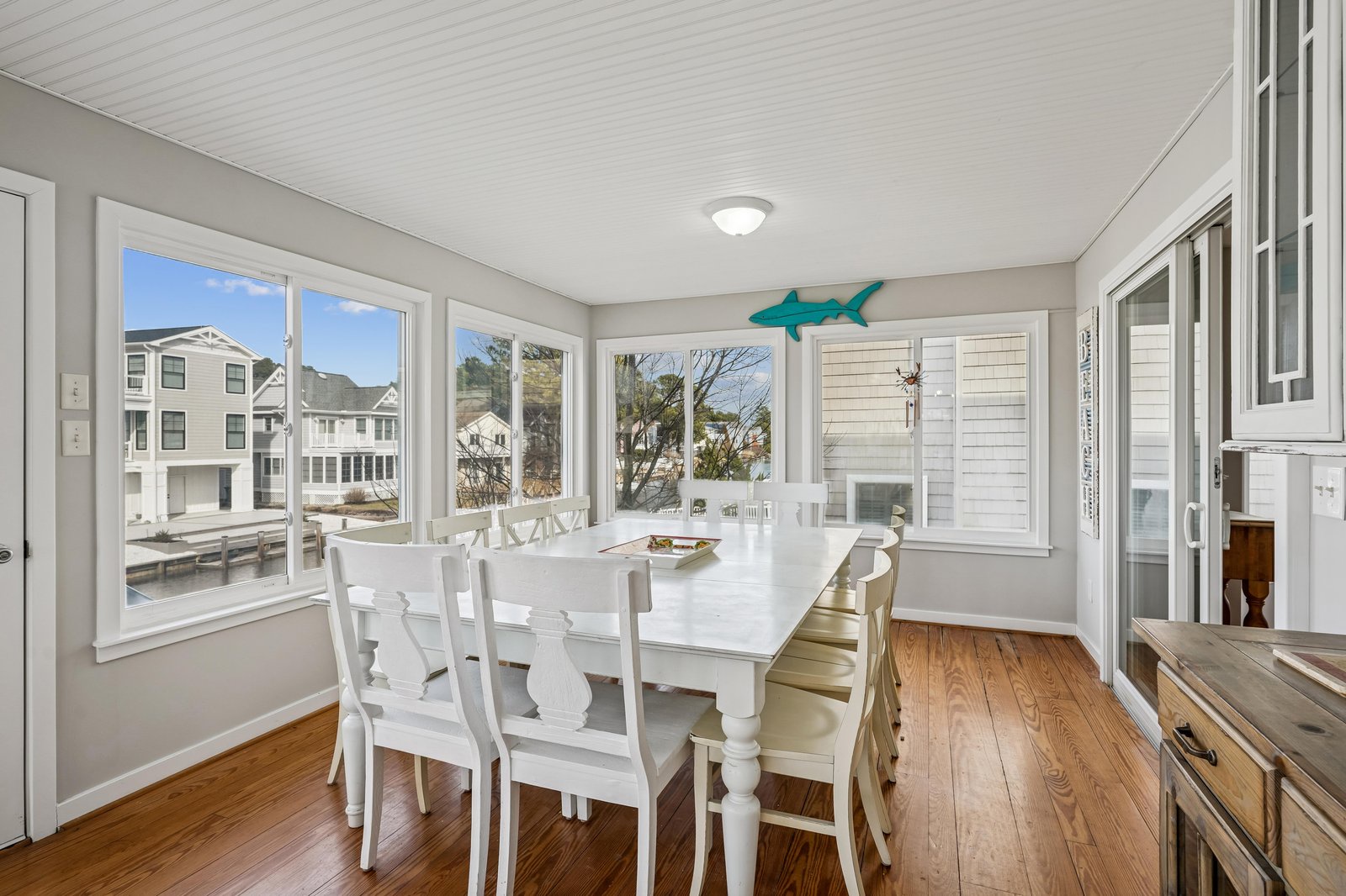 Gather around the elegant white dining table for meals with waterfront views framing your experience. Bright windows, coastal charm, and warm wood floors create a welcoming space.