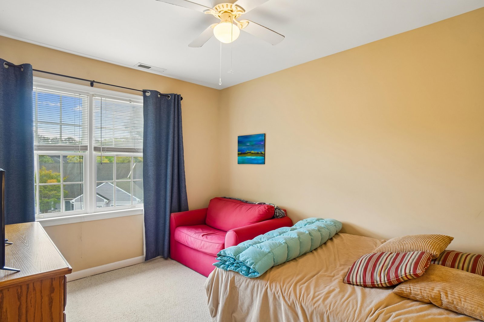 Sink into the plush bed and feel the ceiling fan's gentle breeze as natural light pours through the large window. Your cozy reading nook awaits on the vibrant sofa.