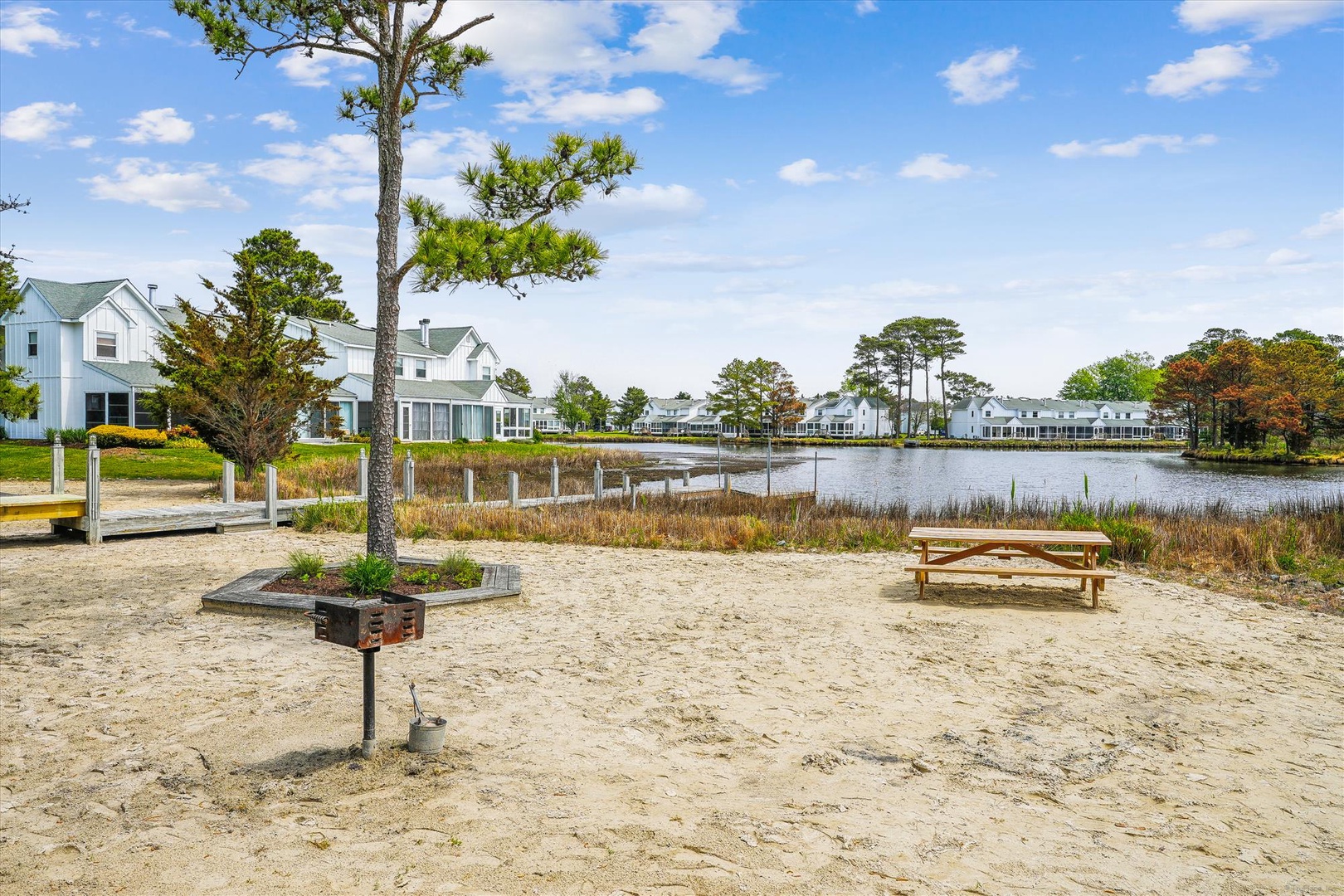 Lakefront community with sandy beach area, picnic tables, and charming vacation homes nestled among mature trees.