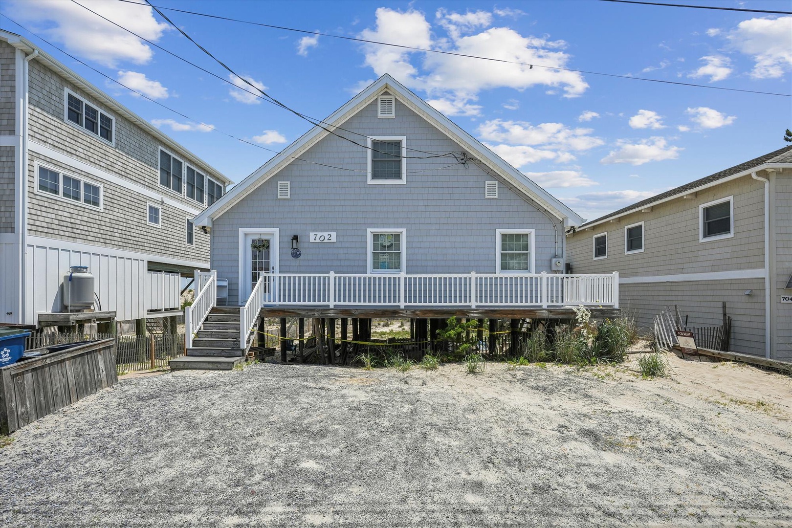 Charming beach cottage at 702, elevated on stilts with coastal character, tucked among neighboring homes in a sandy shore community beneath a bright blue summer sky.