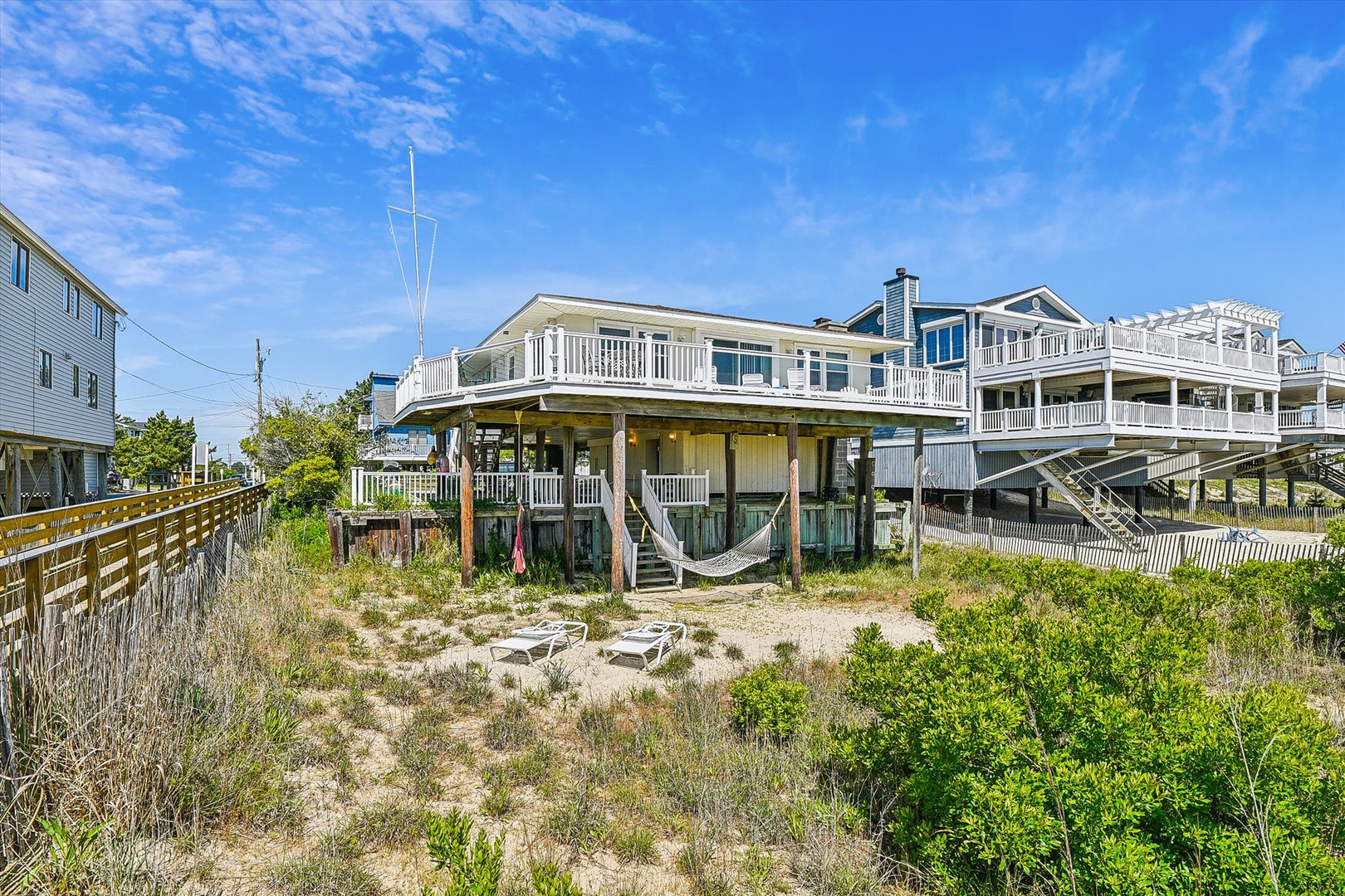Elevated beachfront property featuring expansive wrap-around decks with direct access to sandy dunes and coastal vegetation, positioned in a residential coastal neighborhood under bright skies.