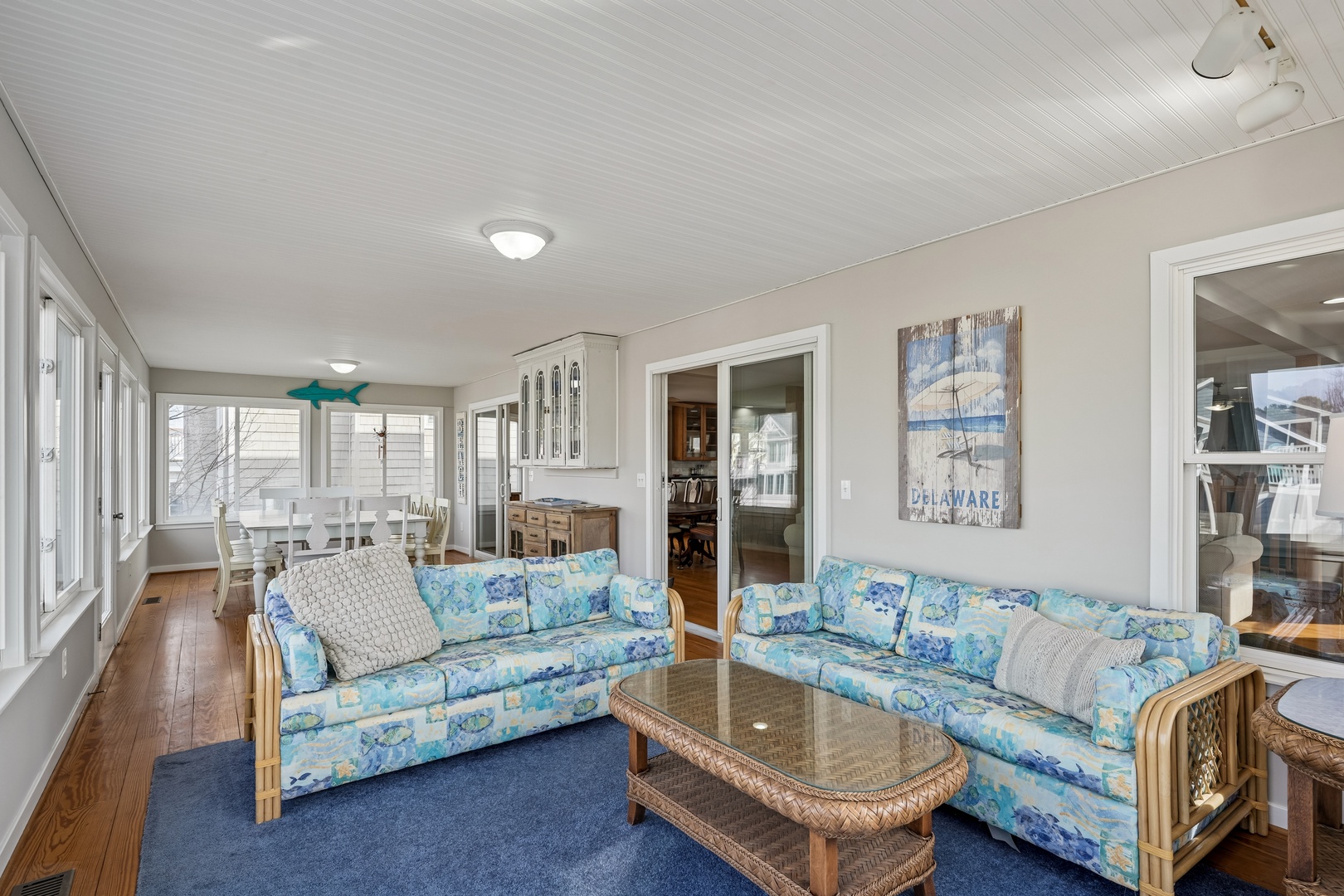 Relax in your bright coastal living room featuring ocean-blue seating, natural wicker furniture, and nautical artwork creating the perfect beach house atmosphere.