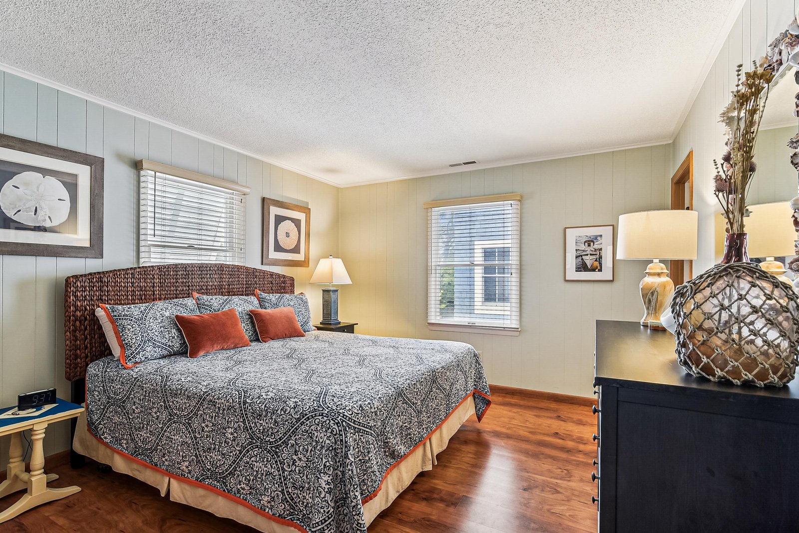 Your coastal-themed bedroom offers a restful retreat with warm wood floors, beachy decor, and soft natural light filtering through the windows.