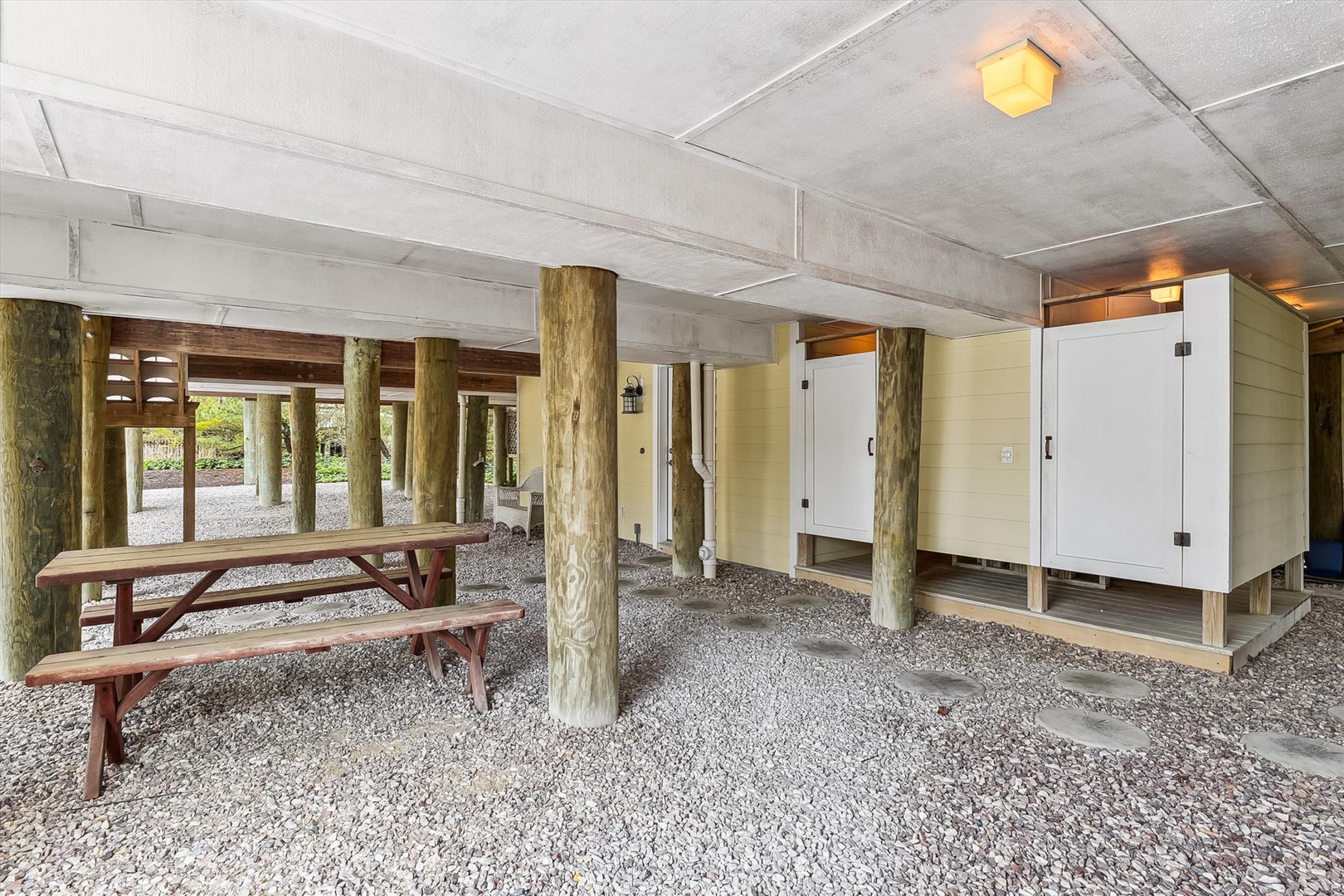 A covered ground-level area with natural wood posts, gravel flooring, a picnic table, and secure storage lockers—perfect for outdoor gear and beach equipment.