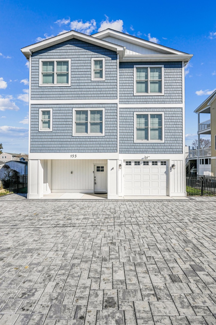 A three-story coastal home with gray shake siding and white trim features generous street-side parking and an attached two-car garage at 155, set under clear blue skies.