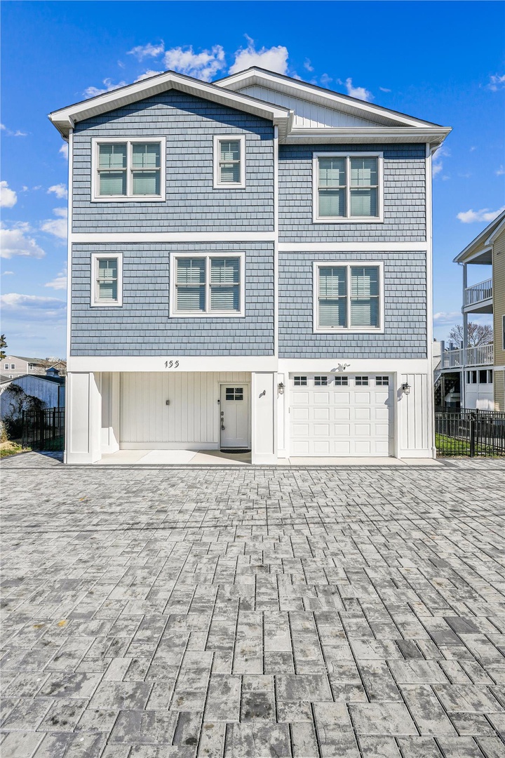 A modern three-story coastal home with blue-gray shingle siding and white trim, featuring a two-car garage and spacious paver-stone driveway at the property entrance.