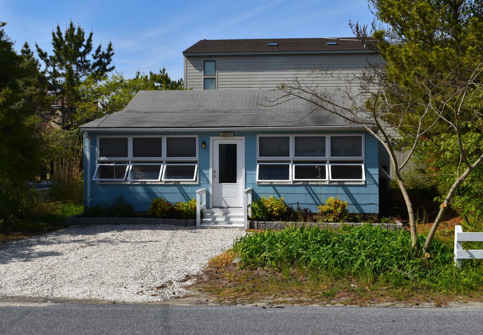 Charming blue beach cottage with gravel driveway and lush greenery, nestled in a quiet coastal neighborhood—perfect for a relaxing seaside getaway.