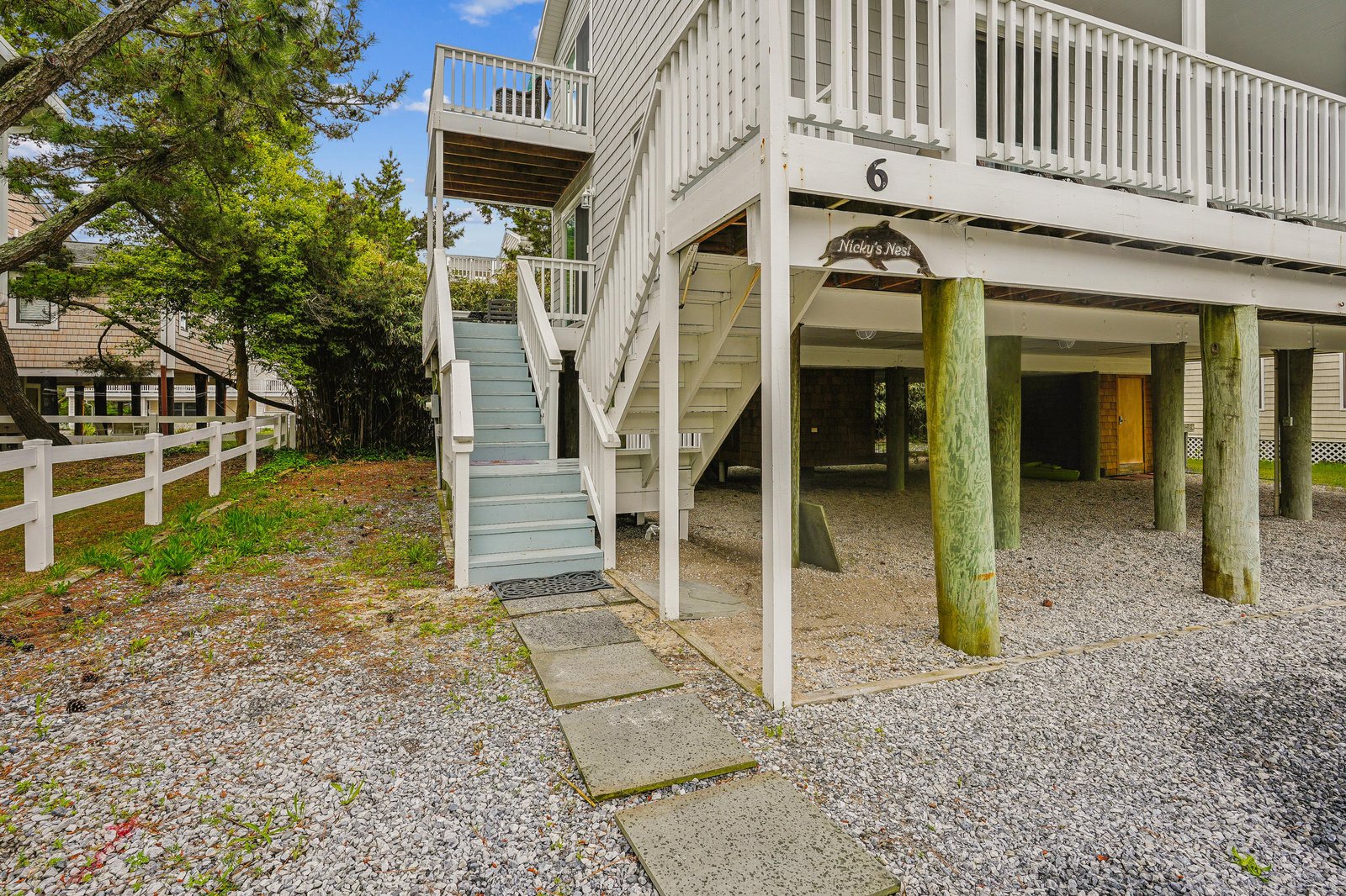 A charming beach cottage elevated on pilings with a covered ground-level parking area and stairway leading to the main entrance, nestled in a coastal neighborhood with mature trees and quiet surroundings.
