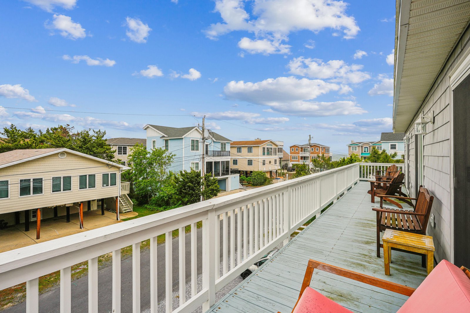 A spacious deck overlooks a quiet residential neighborhood, offering colorful beach cottages and lush greenery under bright coastal skies. Relax in the wooden lounge chairs and soak in the peaceful ambiance of this charming seaside area.