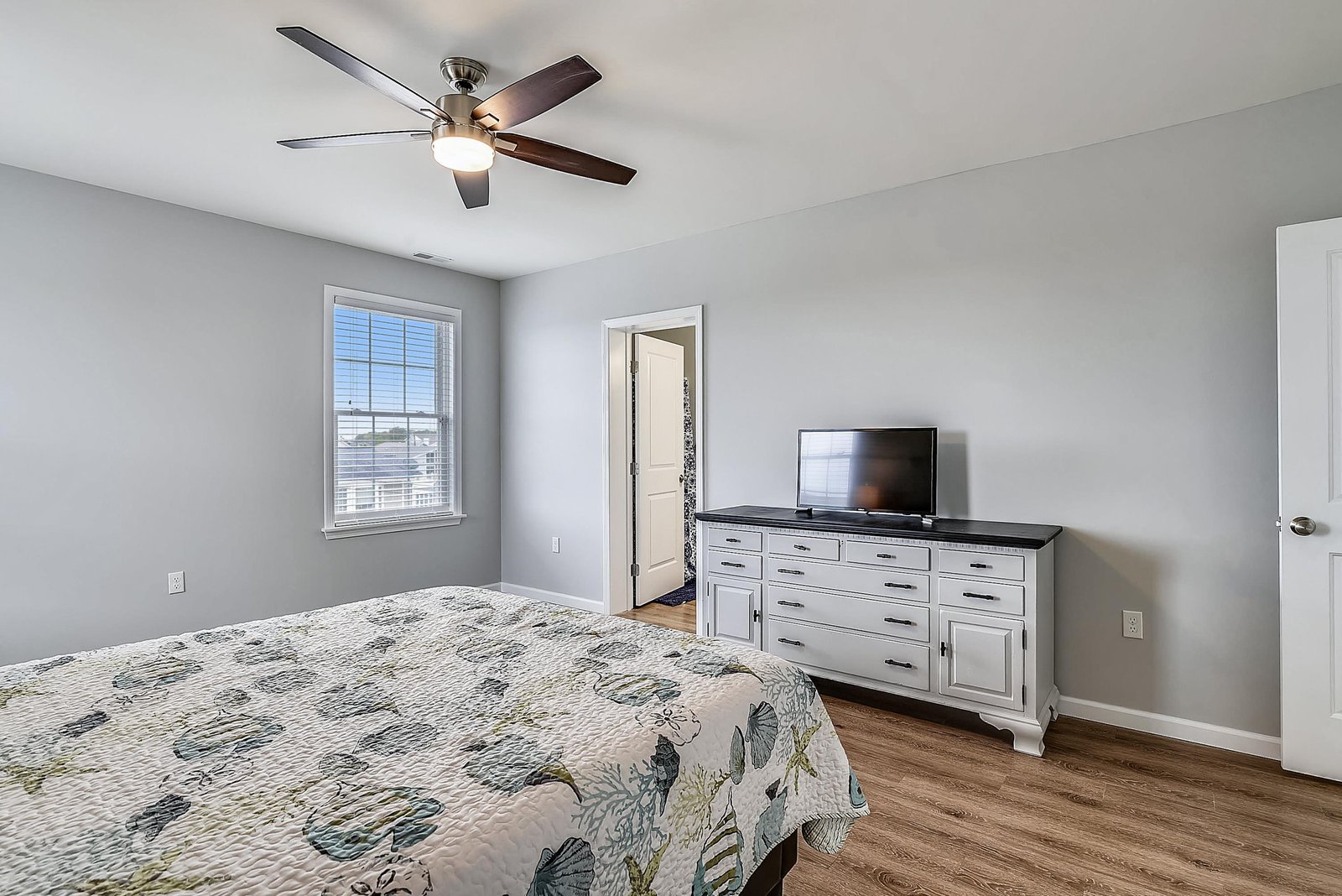 Sink into your peaceful retreat featuring calming gray walls, elegant wood floors, and a stylish ceiling fan overhead. The spacious dresser and flat-screen TV provide all the comforts of home for your stay.