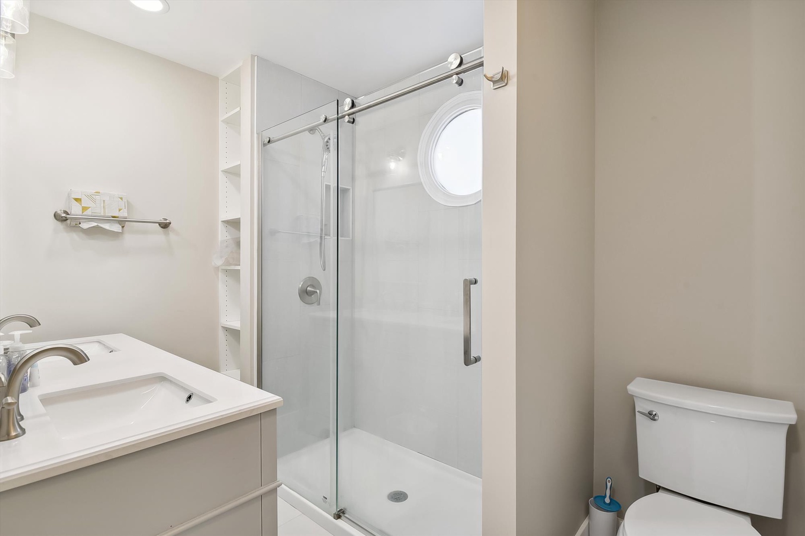Your bright, modern bathroom offers a sleek glass-enclosed shower with porthole window, crisp white fixtures, and thoughtful touches like fluffy towels and convenient open shelving.