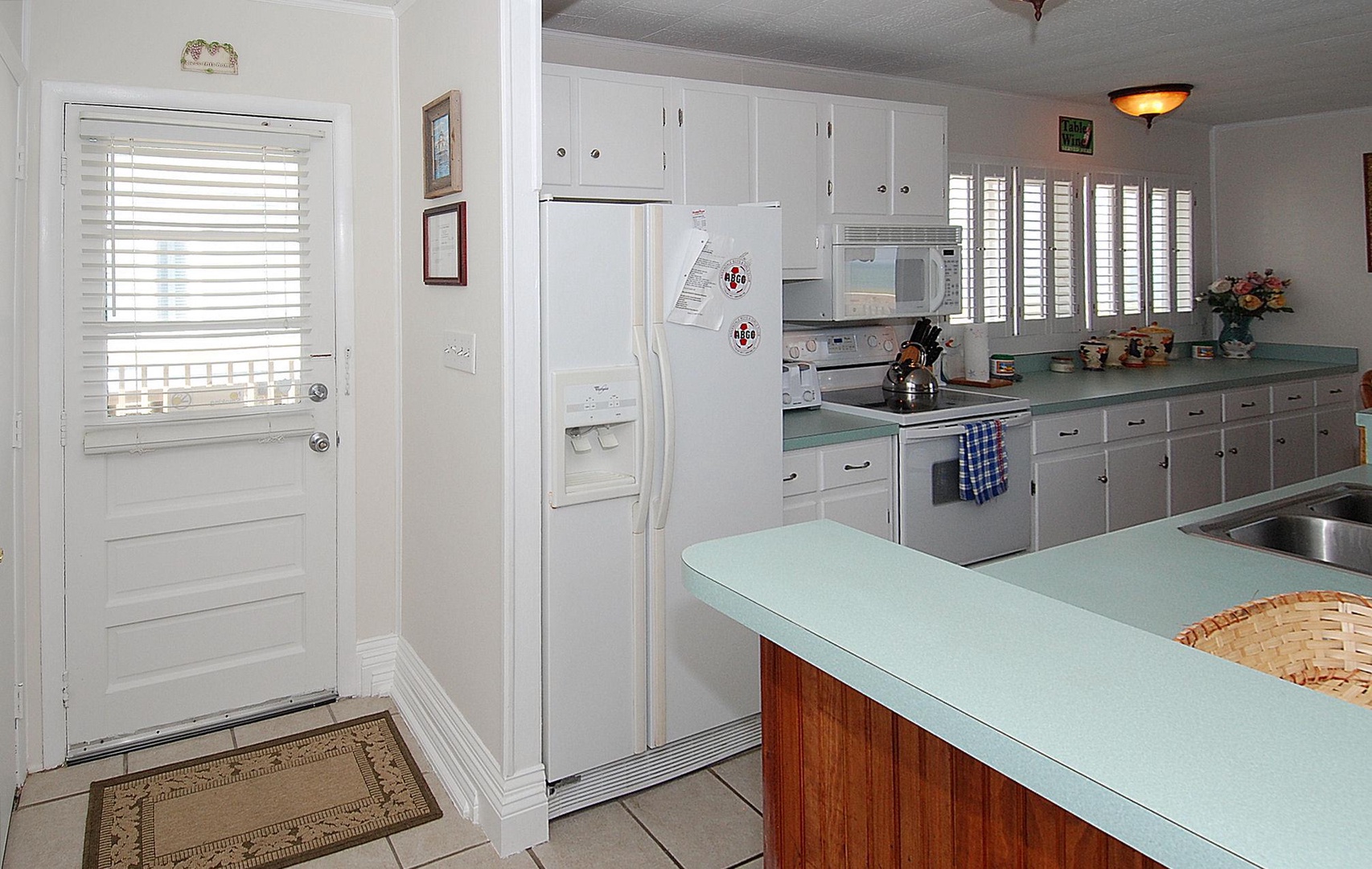 A bright, fully equipped kitchen with mint-green countertops, white cabinetry, and a side-by-side refrigerator. You'll have a microwave, stove, and all the essentials for cooking meals together. The door leads outside, offering easy outdoor access.