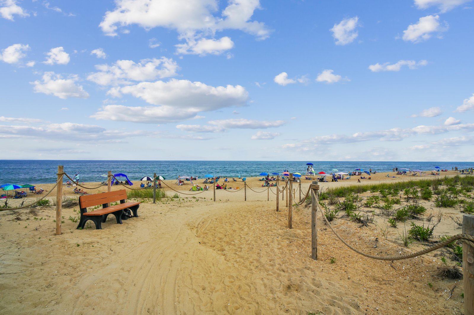 Sandy beach path leads to the sparkling ocean, where colorful umbrellas dot the shore on a beautiful summer day. A bench invites you to pause and soak in the coastal atmosphere.