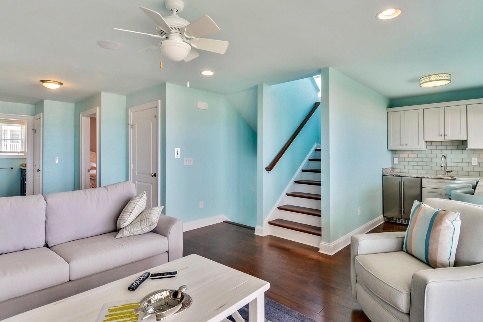 Relax in your coastal-inspired living space with soothing turquoise walls, comfortable seating, and an open layout that flows seamlessly into the modern kitchen.