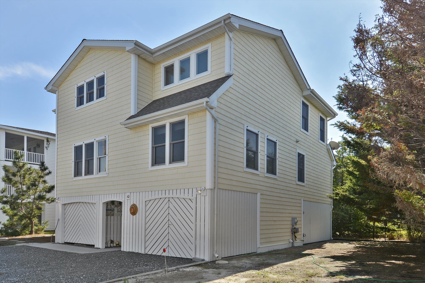Pale yellow two-story rental home with distinctive gabled roofline and ground-level garage. Part of a coastal neighborhood surrounded by mature trees.