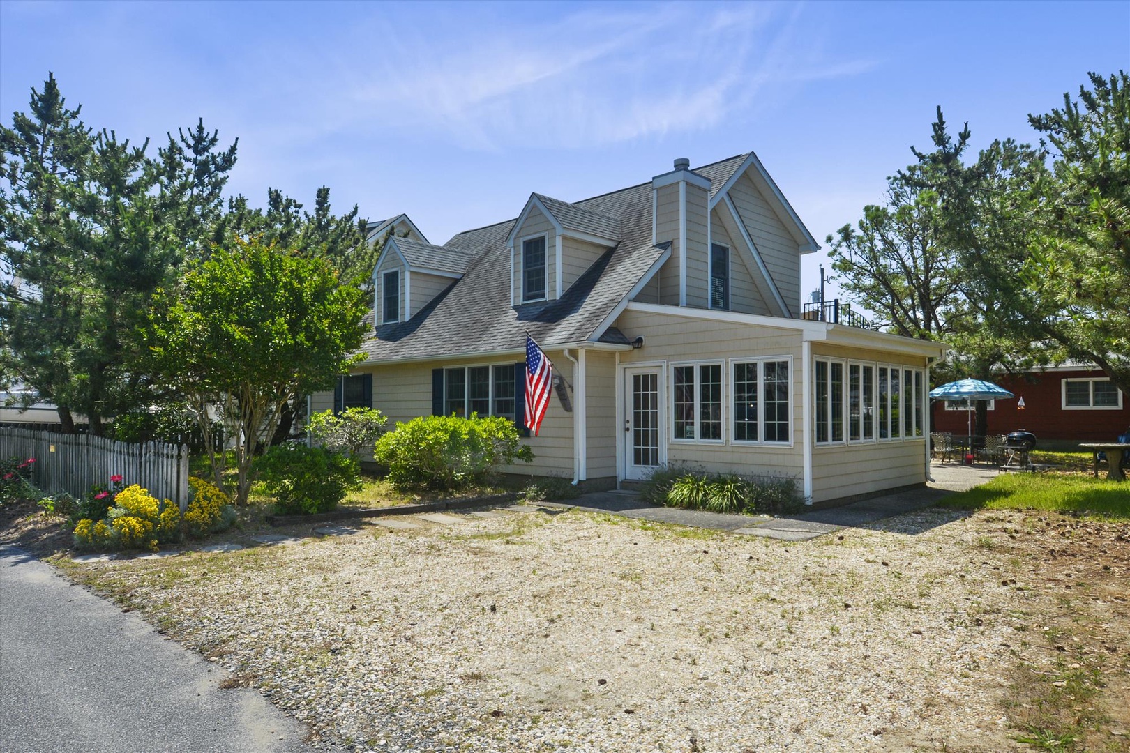 Charming Cape-style vacation home with classic tan siding, welcoming porch, and gravel parking, nestled among lush greenery in a peaceful residential setting.