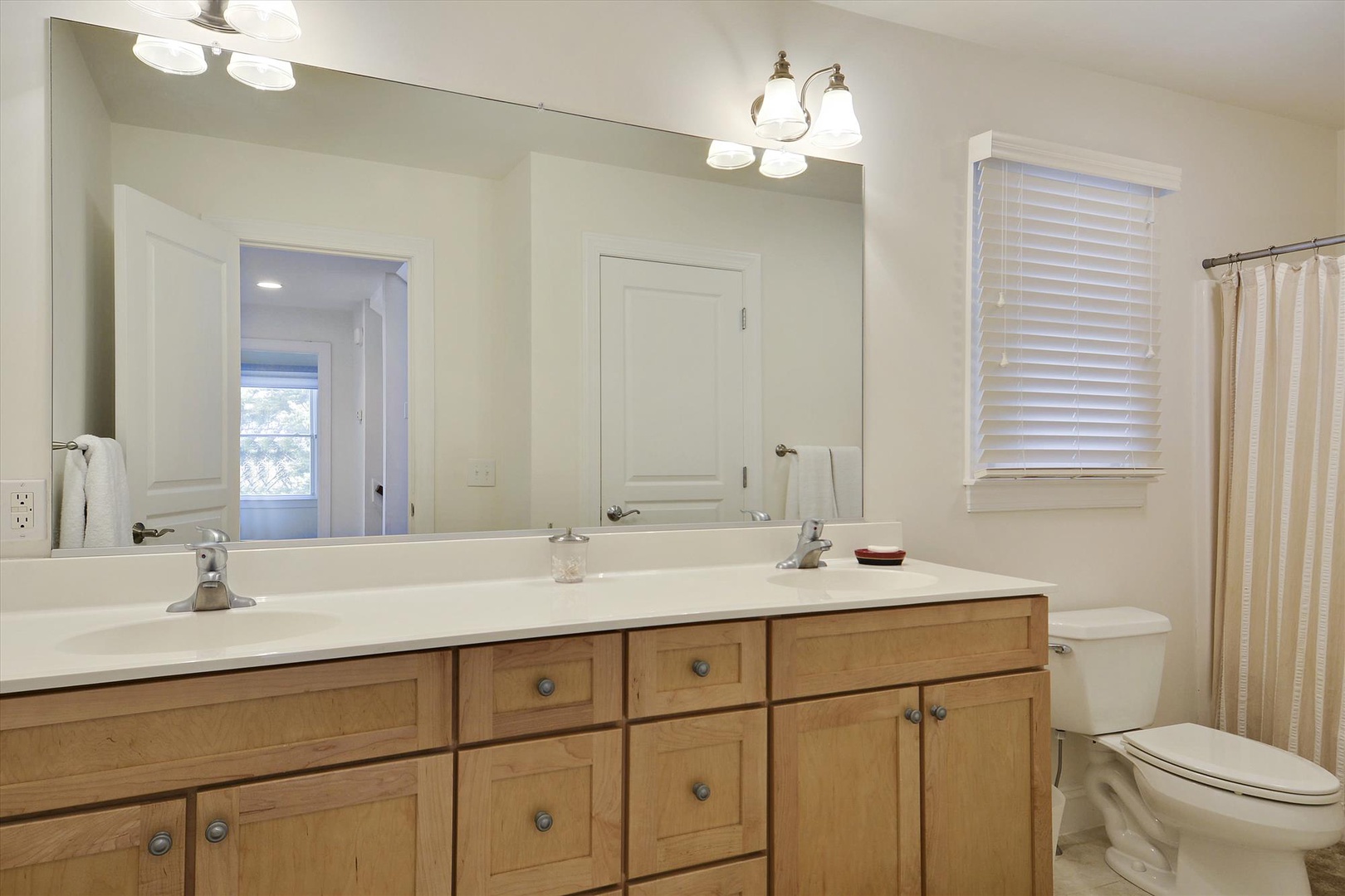 Your spacious bathroom features dual sinks with warm wood cabinetry, a generous mirror reflecting natural light, and a peaceful, bright setting perfect for your morning routine.