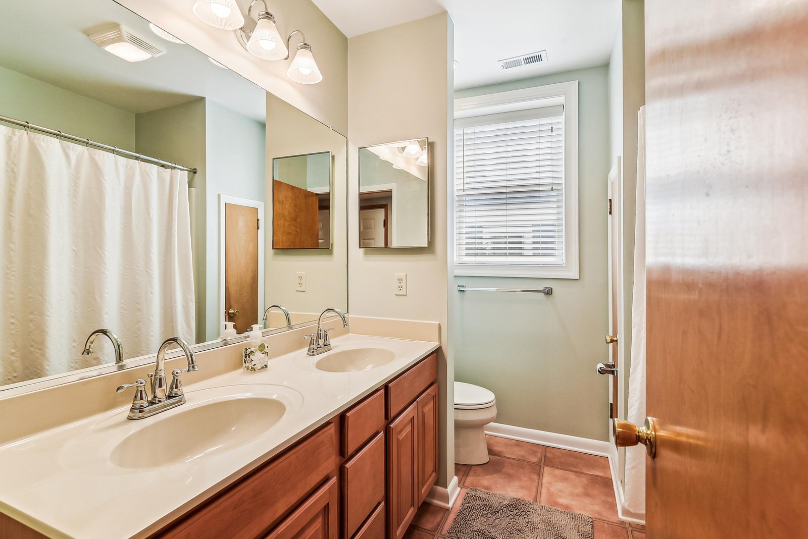 Your private bathroom features dual sinks with warm wood cabinetry, soft lighting, and a bright window that fills the space with natural light—perfect for your morning routine.
