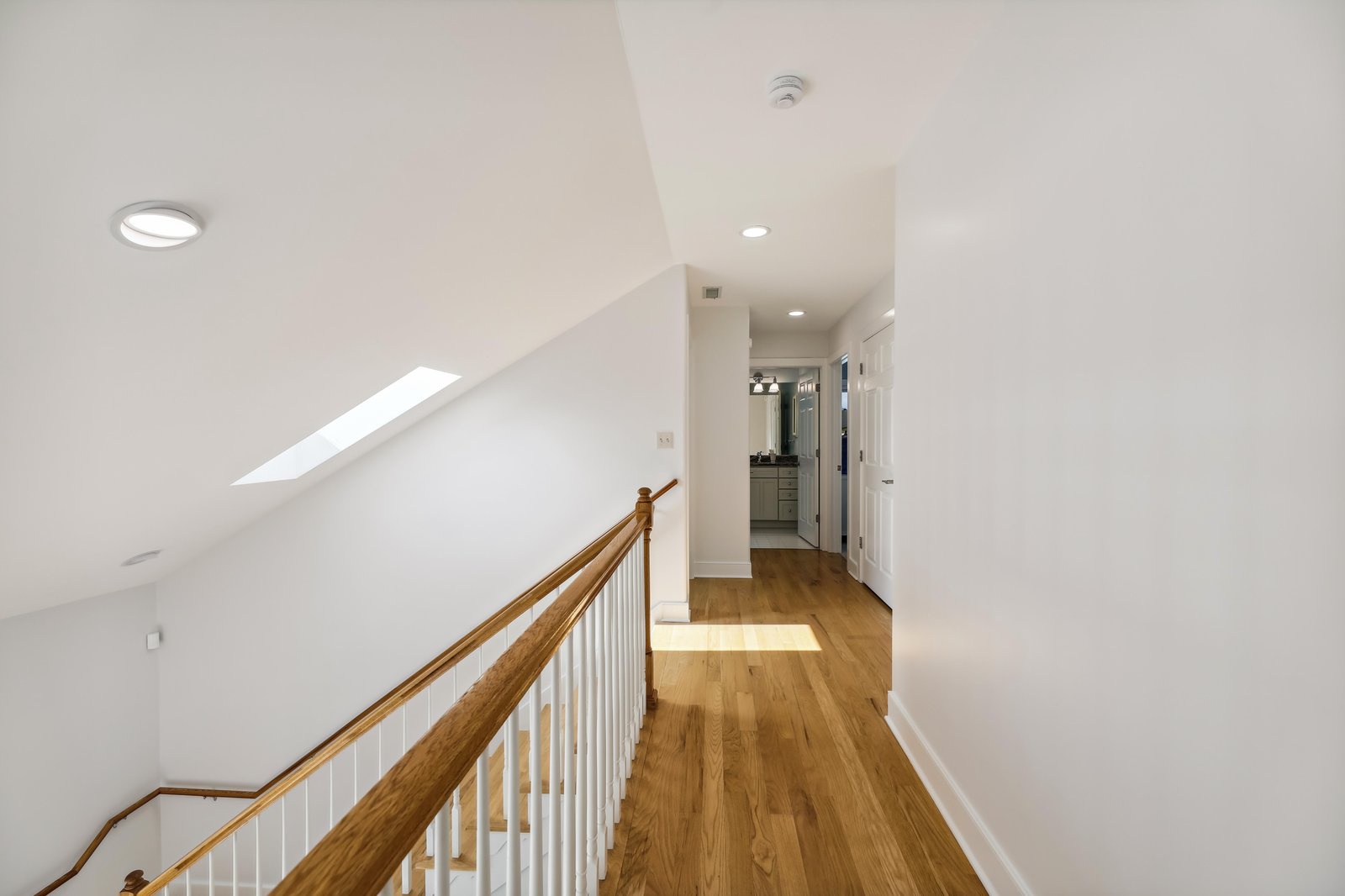 Step into your bright upper-level hallway, where natural light from the skylight warms gleaming hardwood floors and leads you to spacious rooms ahead.