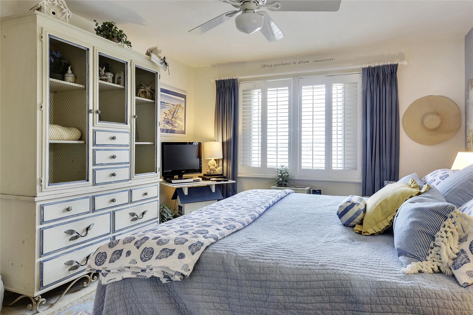 Sink into the plush queen bed surrounded by coastal charm, where soft linens and gentle ceiling fan breezes promise restful nights. Your wardrobe fits perfectly in the elegant armoire, while sunlight filters through plantation shutters.