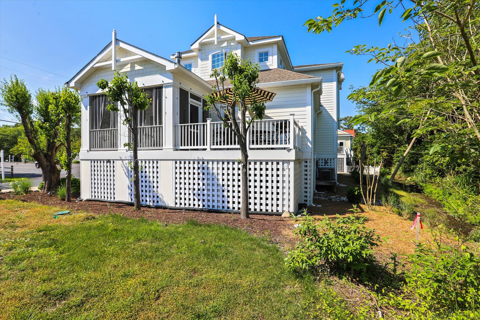 A charming two-story property featuring classic white siding, a welcoming screened porch, and lush garden surroundings—nestled in a peaceful residential area with abundant natural greenery and clear blue skies overhead.