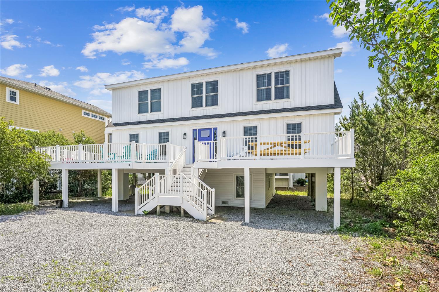 Charming coastal home with blue front door and spacious covered parking. Elevated design with white railings and mature landscaping creates perfect beach getaway setting.
