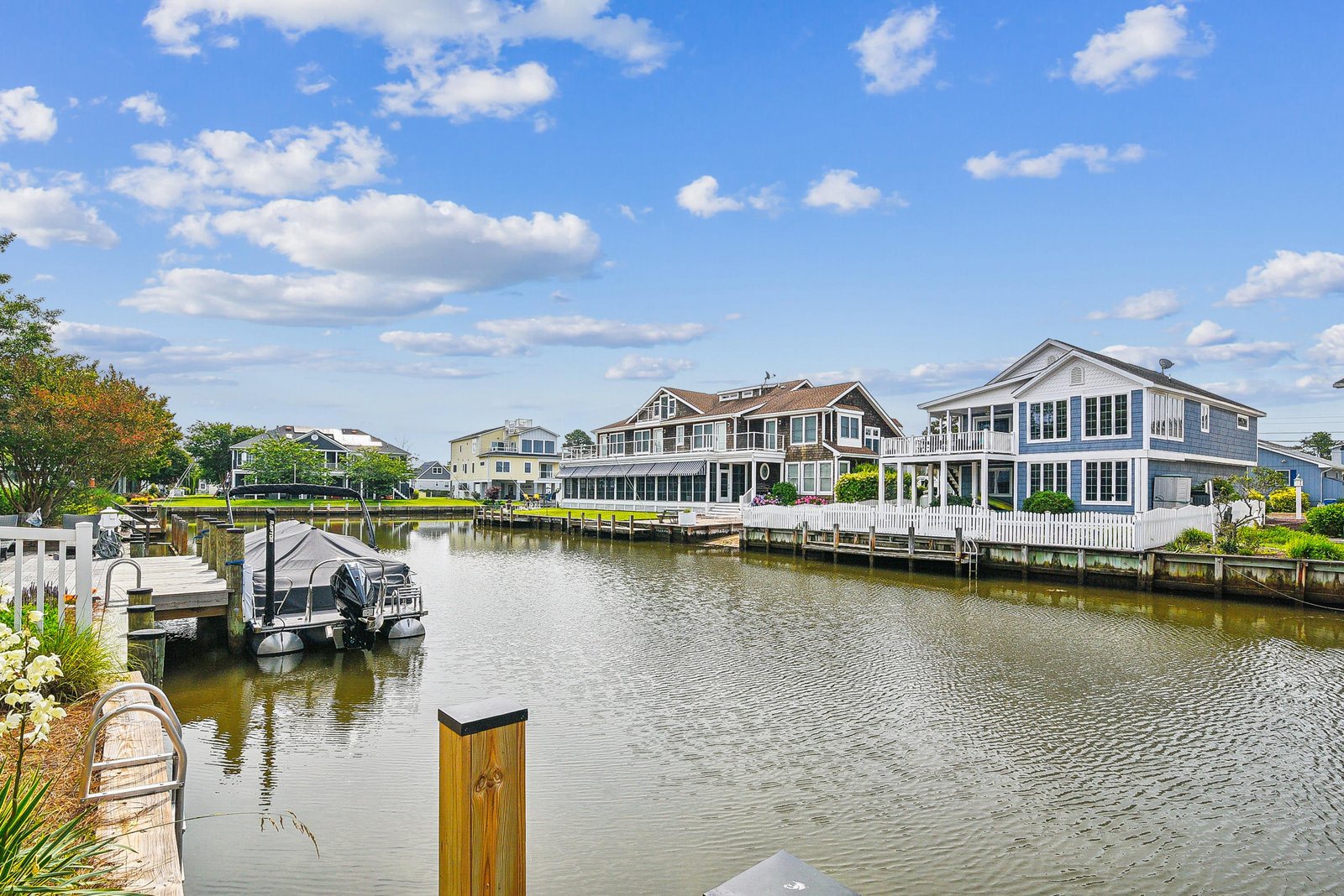 A tranquil waterfront neighborhood features charming homes lining a calm canal with private docks and boats, framed by blue skies and lush greenery.
