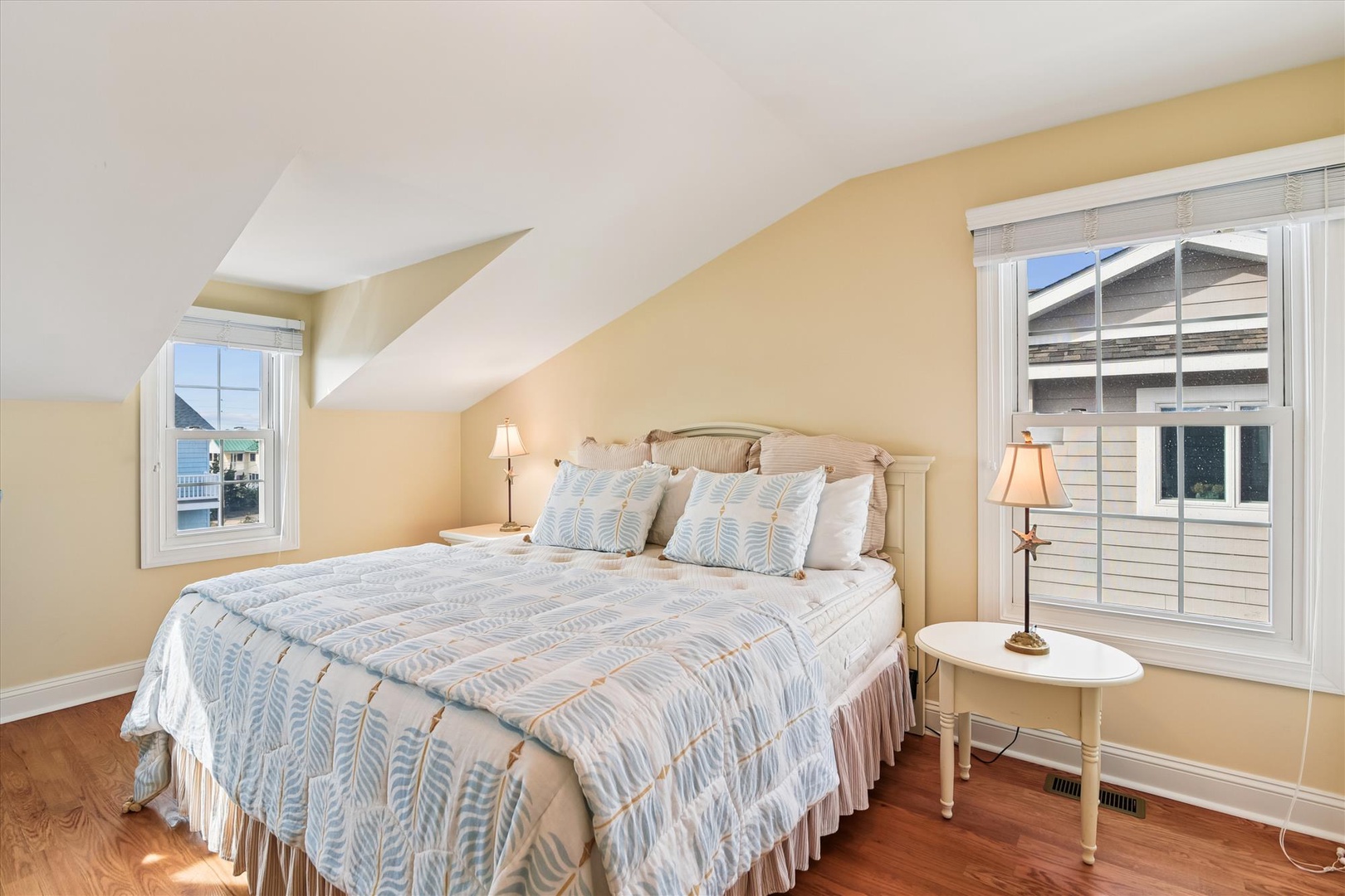 Your bright, cozy bedroom features a welcoming king bed with plush linens, warm yellow walls, and lovely natural light from two large windows—perfect for restful mornings.
