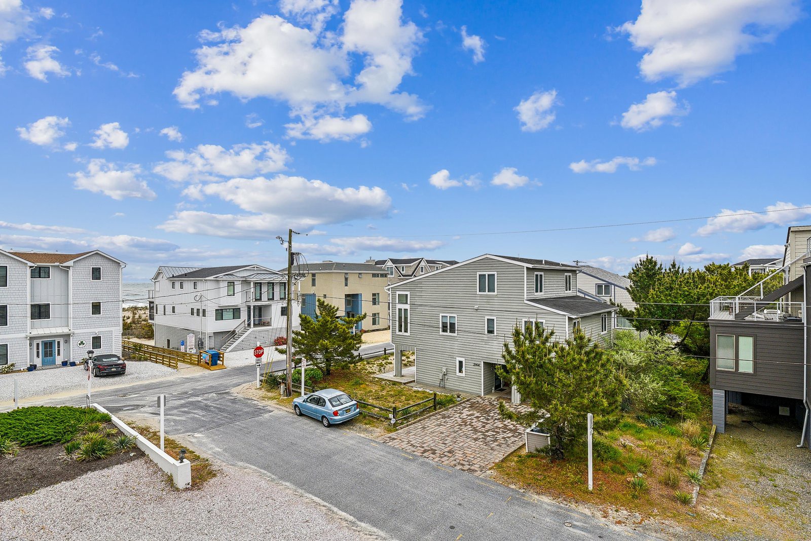 A coastal neighborhood with beach houses, parking areas, and quiet residential streets beneath a bright blue sky with scattered clouds.
