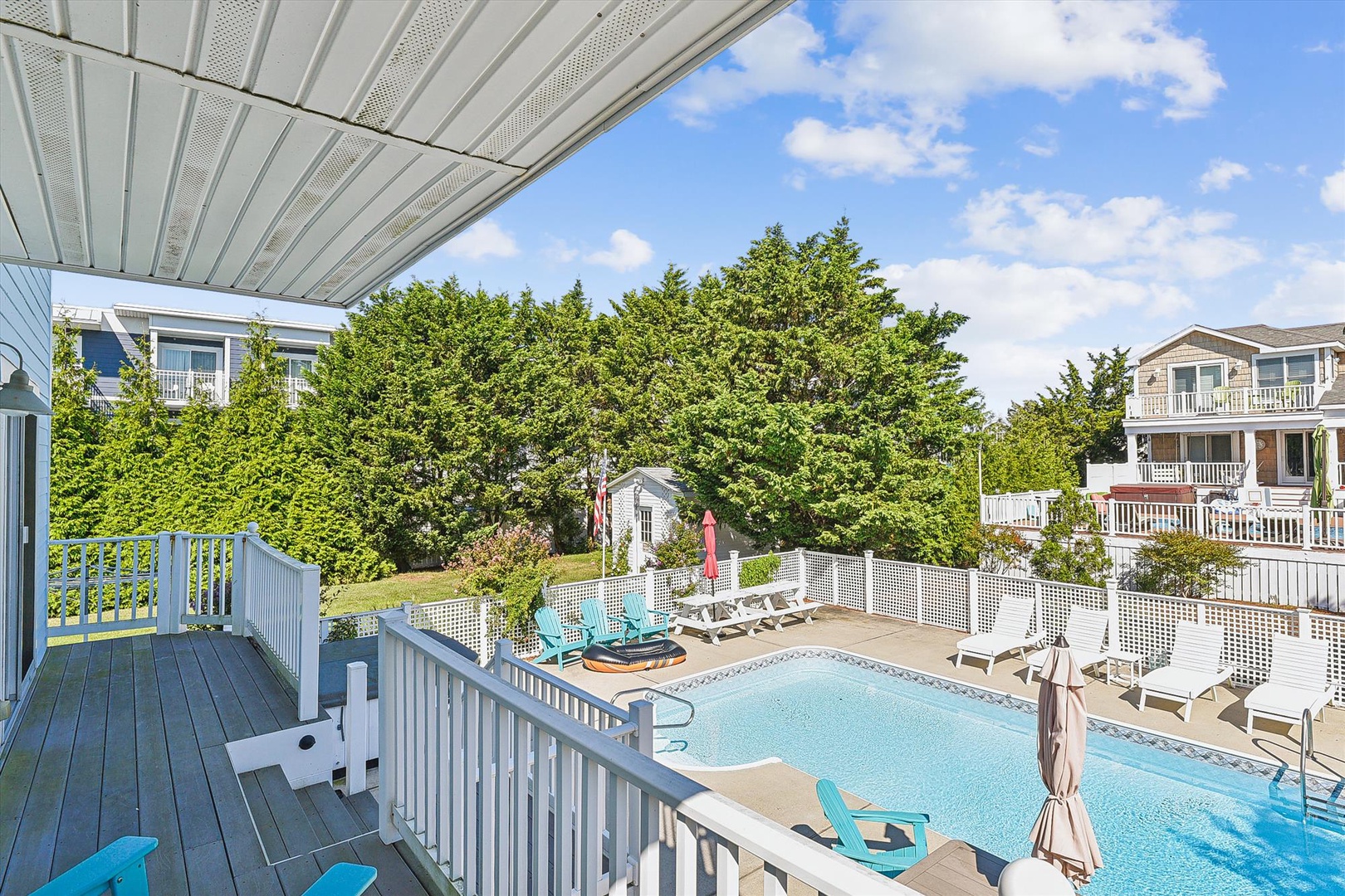 Property features a private pool with lounge chairs, turquoise accent seating, and a covered second-floor deck overlooking a mature, tree-lined neighborhood with nearby coastal homes.