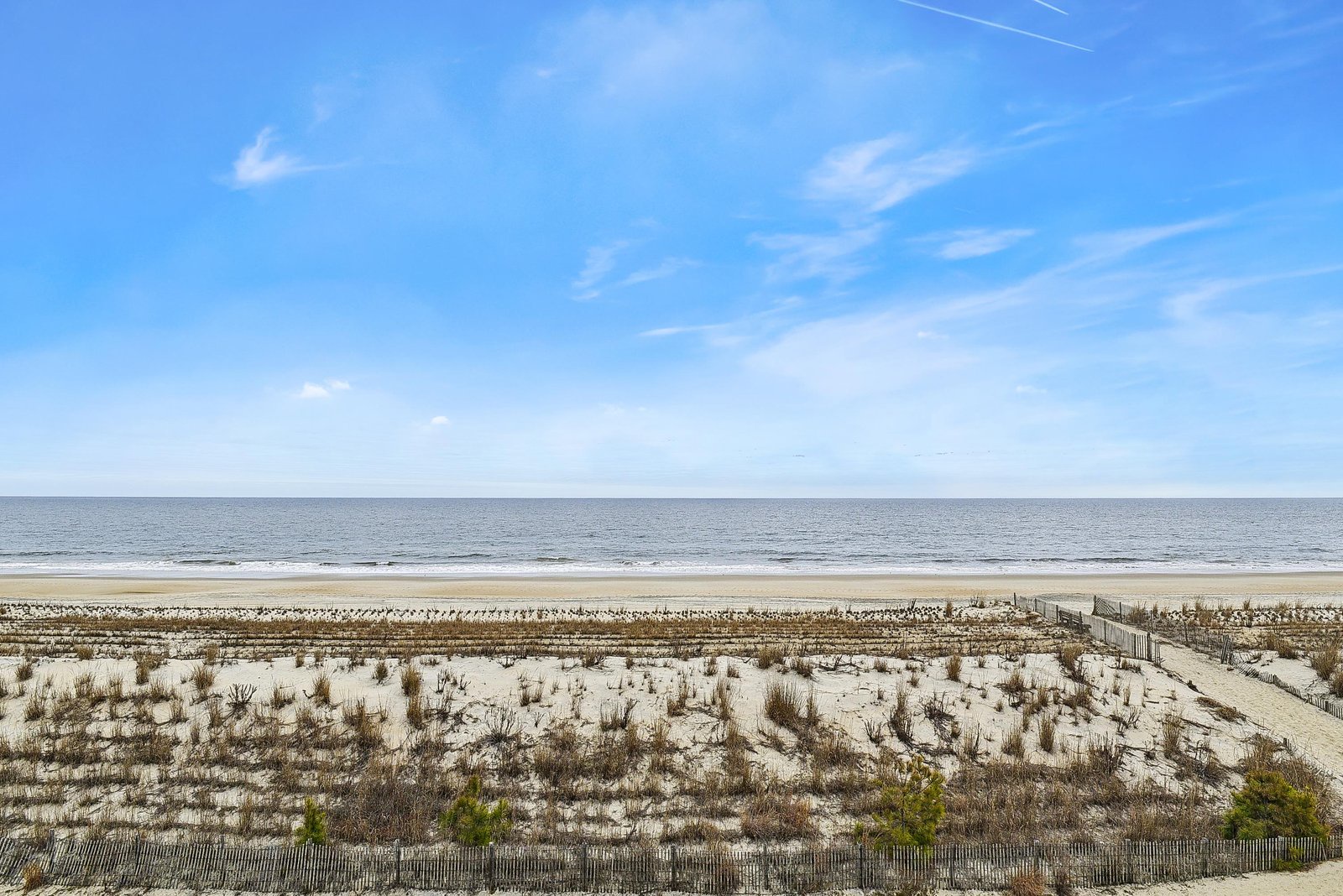 Expansive sandy beach meets the open sea under clear blue skies, with natural dune grasses and a peaceful stretch of shoreline defining this coastal property's surroundings.