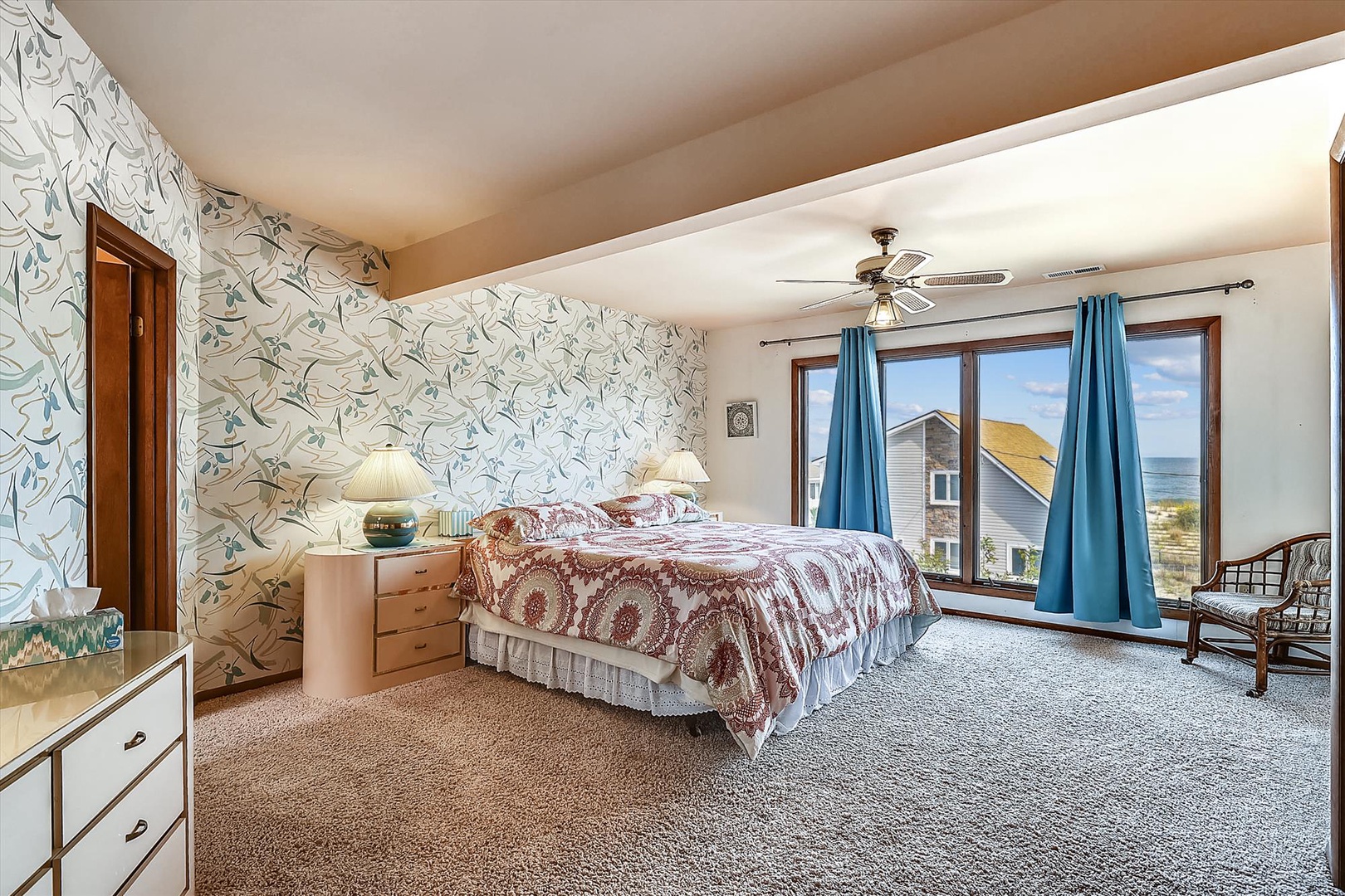 Unwind in your spacious bedroom featuring charming bird-themed wallpaper, a plush bed, and ocean breezes through large windows with peaceful neighborhood views.