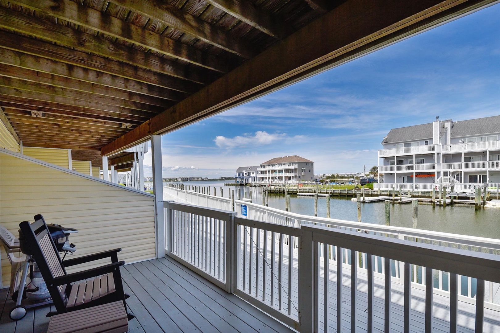 Waterfront seating area with marina and coastal building views, situated under covered wooden ceiling and white railing deck with chairs overlooking calm waters.