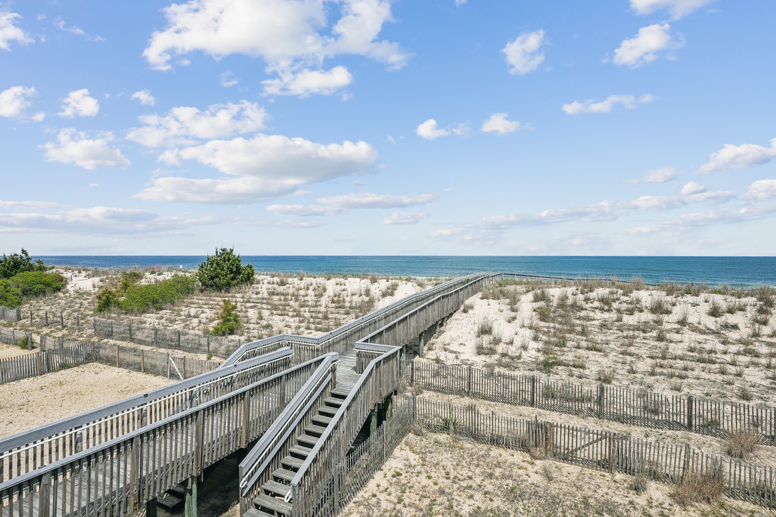 A wooden boardwalk winds through protected dunes, leading to pristine oceanfront access where the blue Atlantic meets sandy shores beneath an open coastal sky.