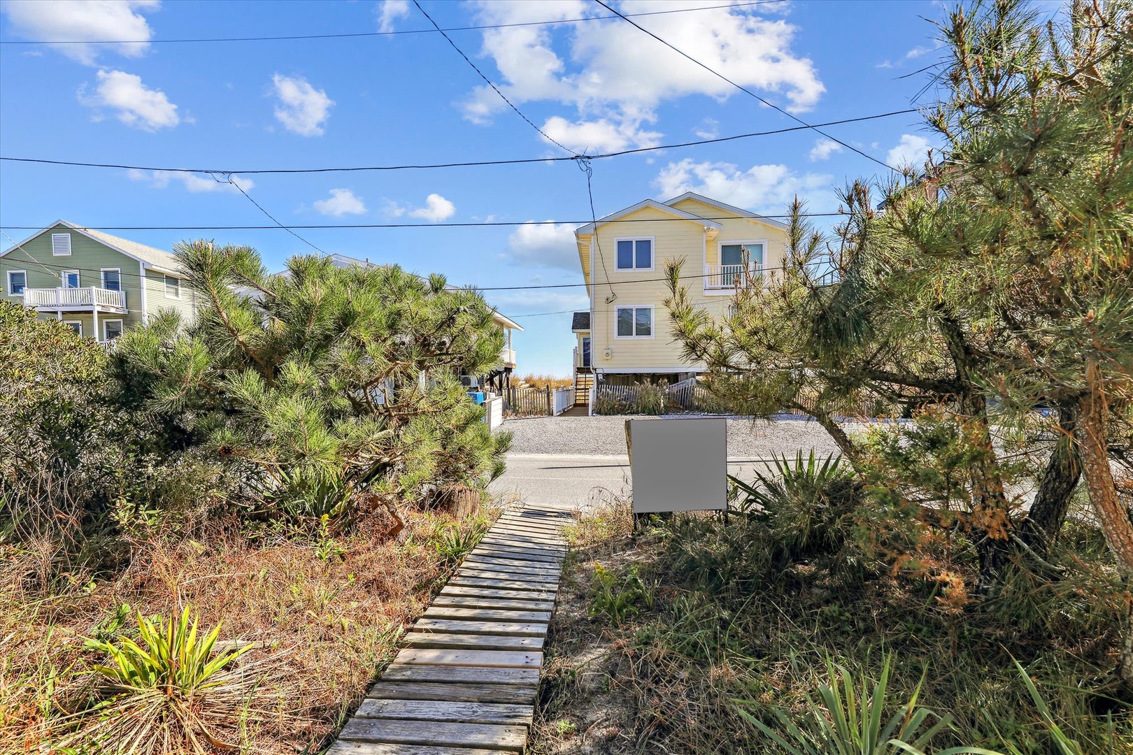 A charming beachside property nestled among coastal pines and native landscaping, with a welcoming boardwalk leading to the entrance. The home features elevated coastal architecture and sits within a peaceful residential neighborhood, just steps from the 