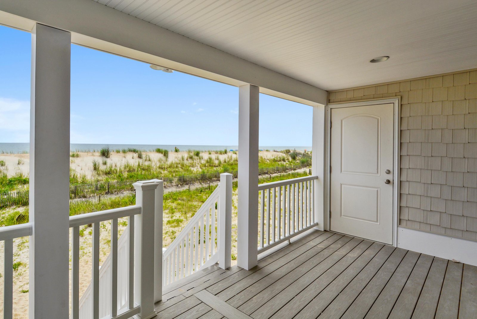The property's covered entrance opens to a serene coastal landscape with sandy dunes and beach grasses just beyond, offering a peaceful seaside arrival.