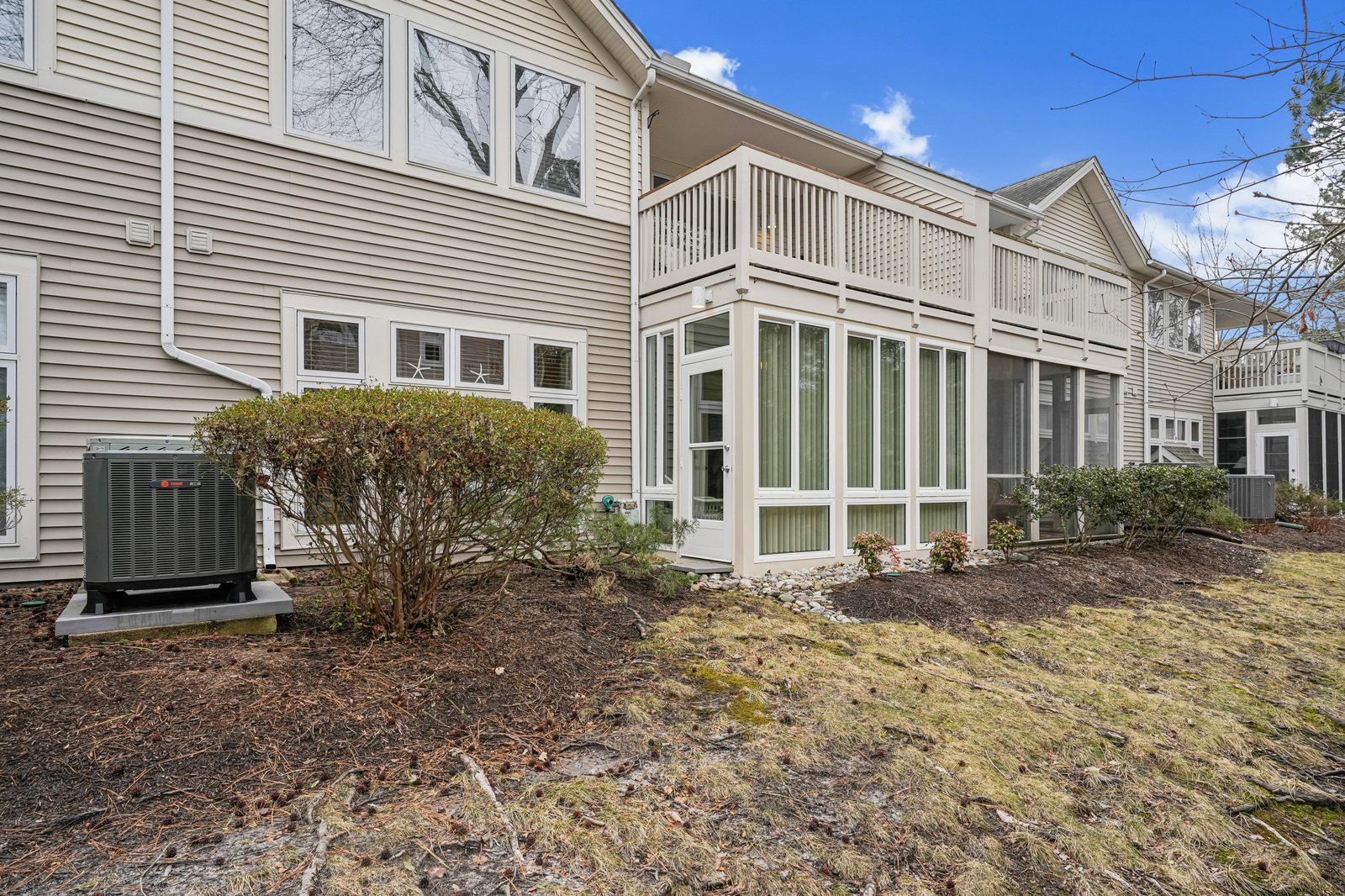 A well-maintained multi-unit property featuring light tan siding, multiple balconies, and a landscaped yard with evergreen shrubs, captured under clear blue skies in early spring.