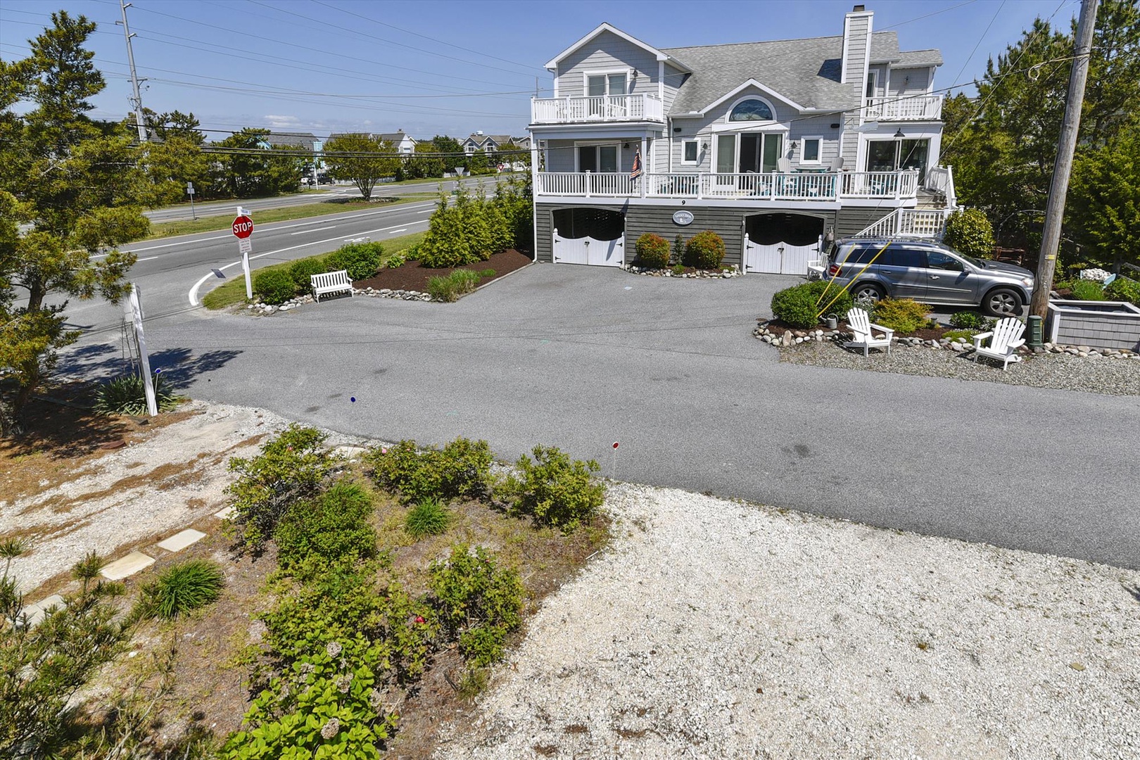 Elevated beach house with spacious driveway, multiple balconies, and white Adirondack chairs for enjoying coastal breezes and nearby ocean views.