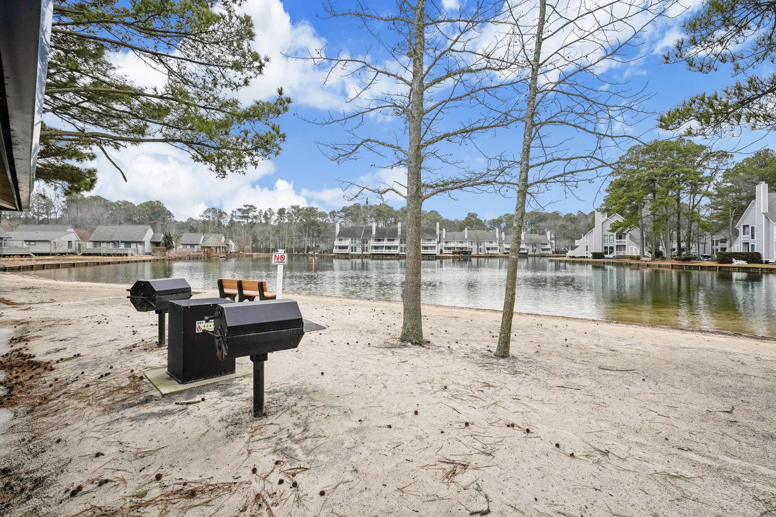 Lakefront community with sandy beach area, BBQ grills, and peaceful water views surrounded by residential buildings.