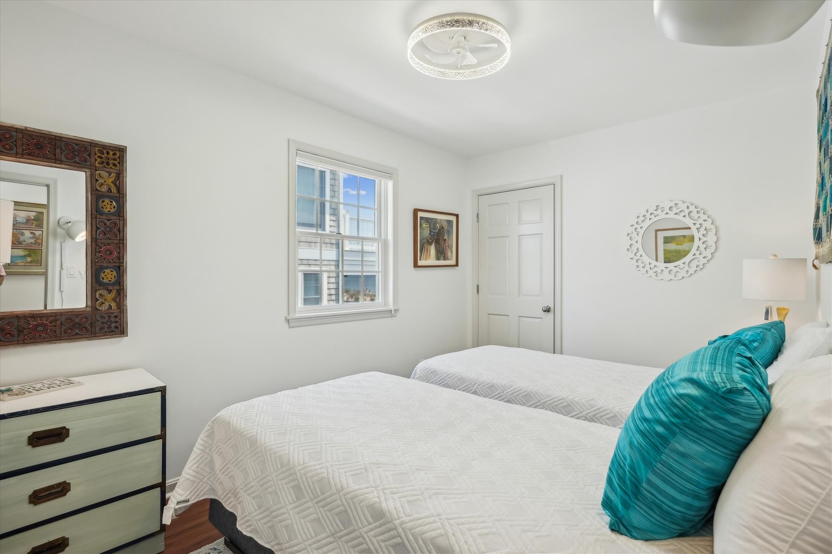 Crisp white walls and plush bedding create your serene retreat. Natural light pours through the window, highlighting thoughtful décor touches and a pop of turquoise for coastal charm.