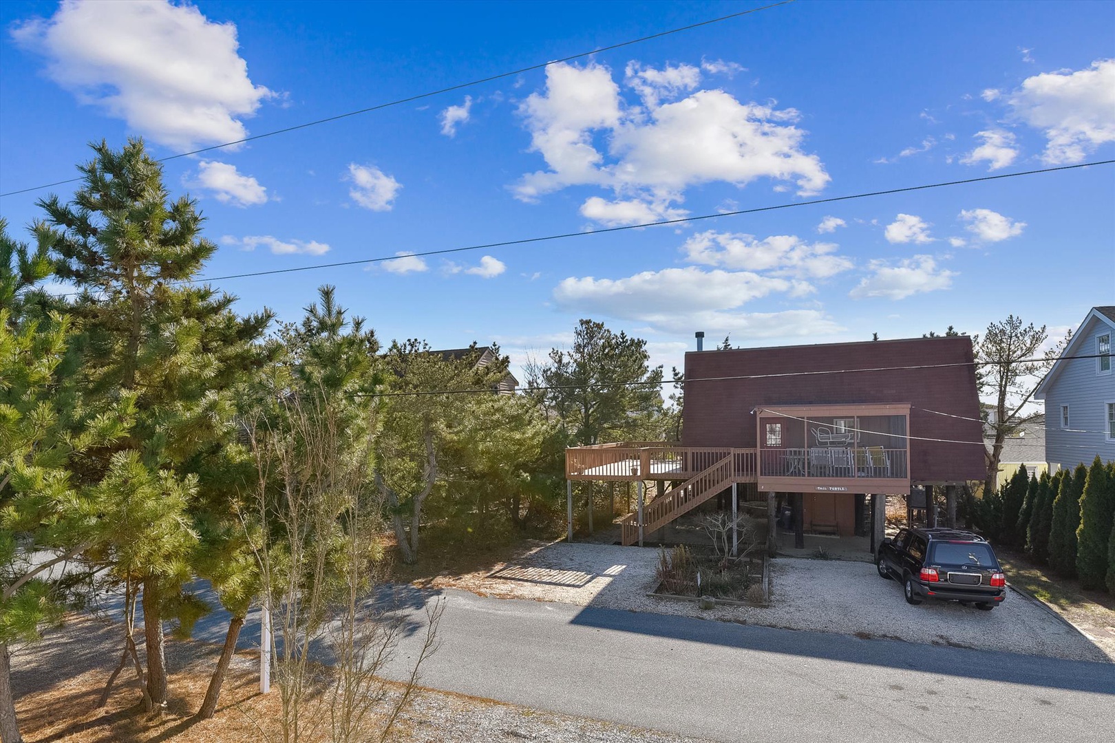 Elevated two-story vacation home surrounded by mature pines and natural landscaping, featuring an upper deck and covered parking area in a peaceful wooded setting.