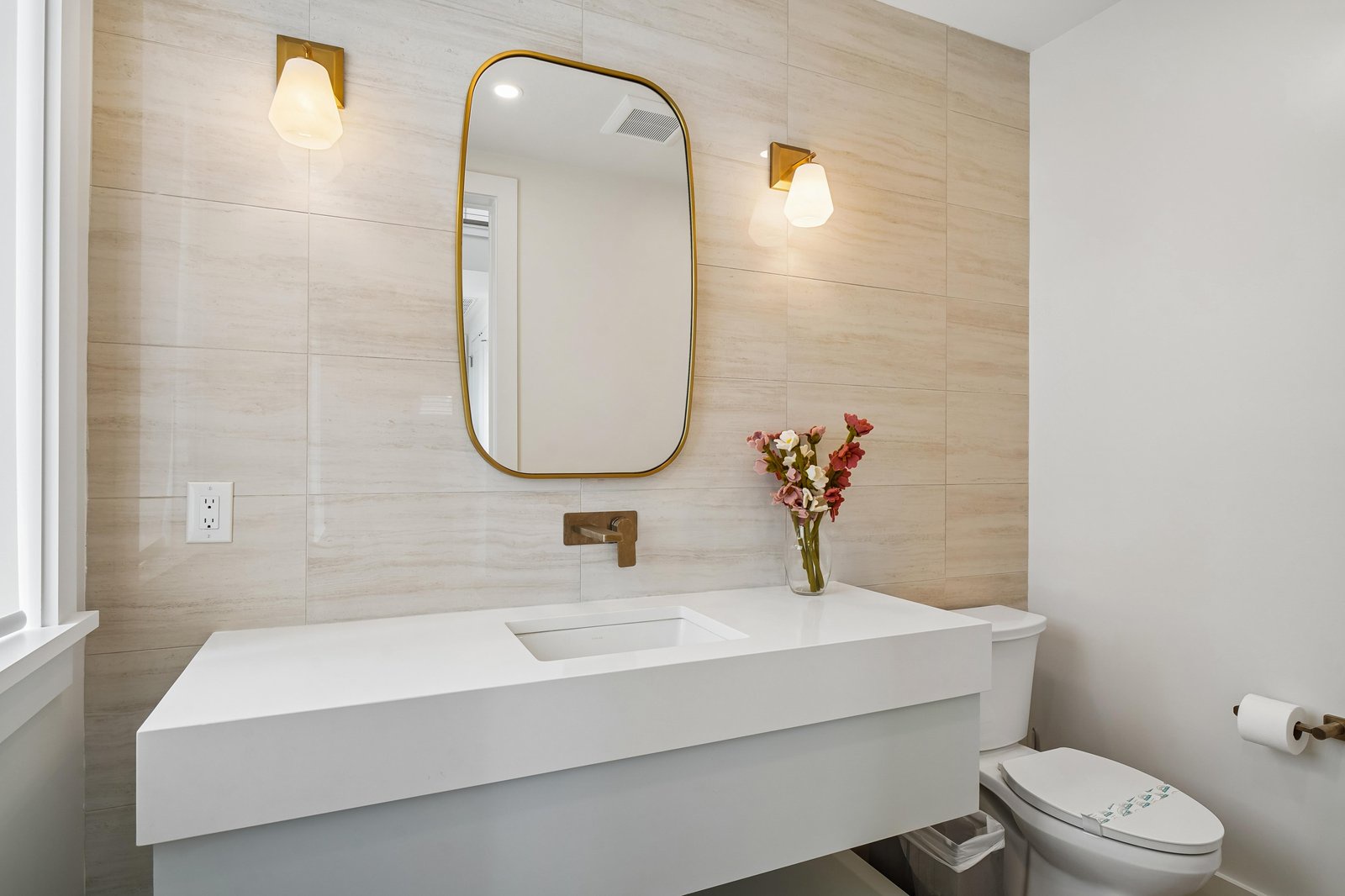 Relax in this bright, modern bathroom with sleek white counters, warm wood-textured walls, and brass accents. Fresh flowers and elegant sconces complete the refined, calming space.