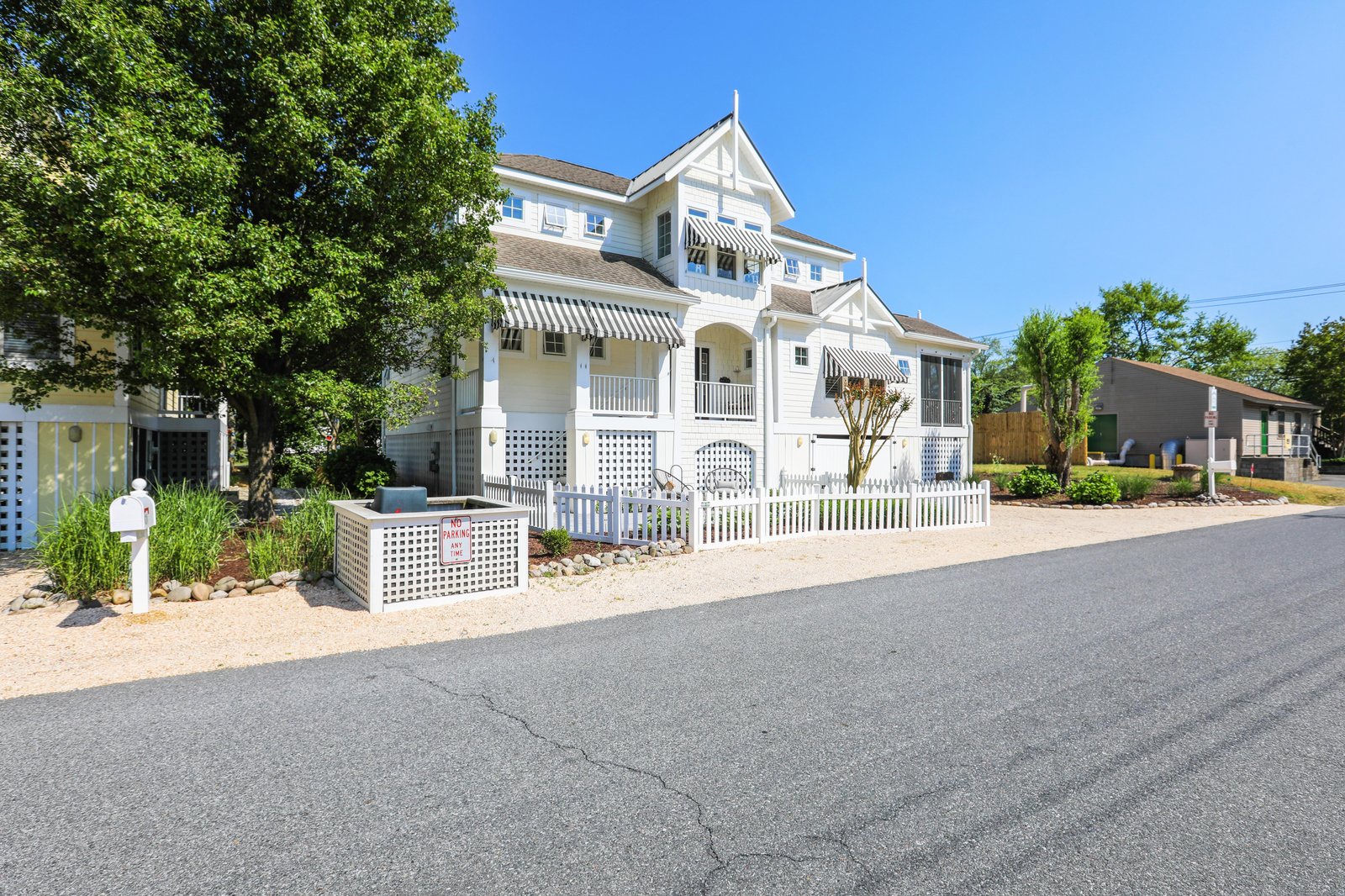 A charming white beach cottage with striped awnings and decorative lattice fencing welcomes guests in a tree-lined neighborhood on a sunny day.