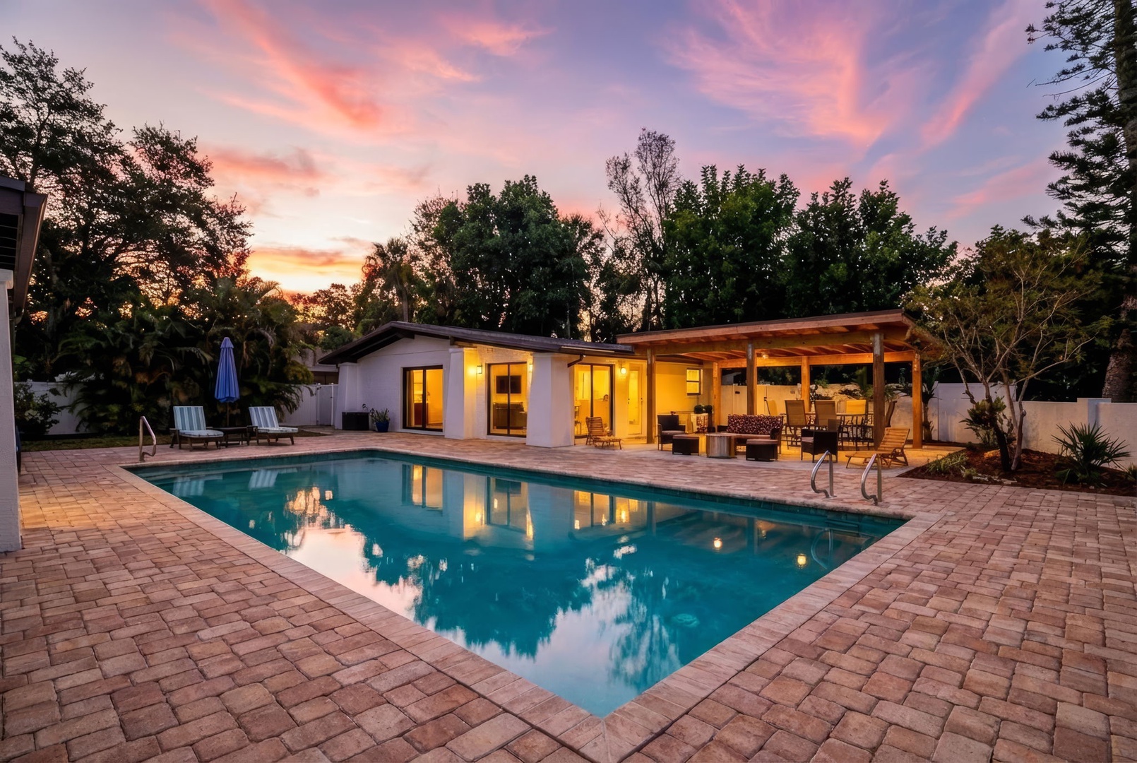 Modern property with pool and covered outdoor dining area surrounded by mature landscaping at sunset.