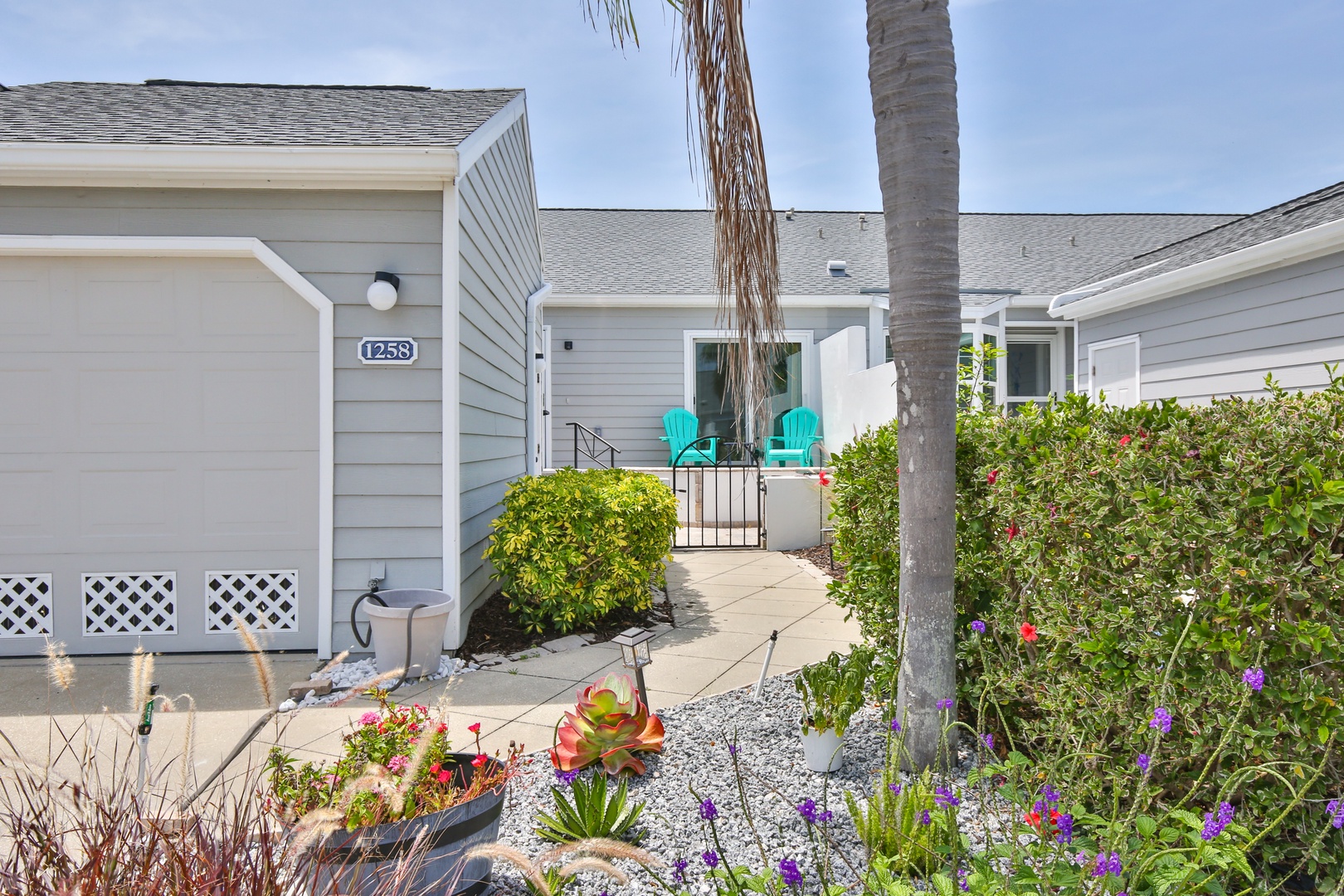 A charming coastal property entrance with inviting turquoise chairs, lush garden landscaping featuring colorful flowers and succulents, and a peaceful pathway that leads to your vacation retreat.