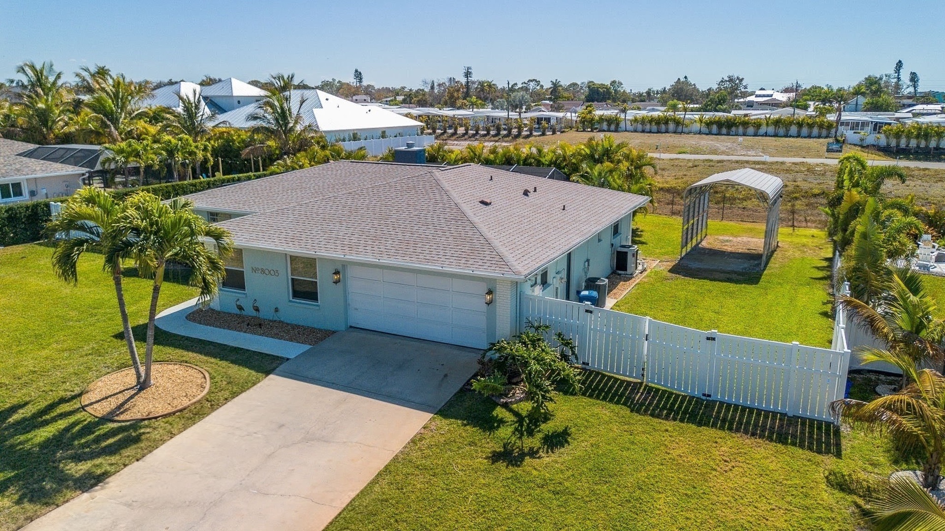 Aerial view of a charming single-story home with tropical landscaping and covered parking in a peaceful residential neighborhood.