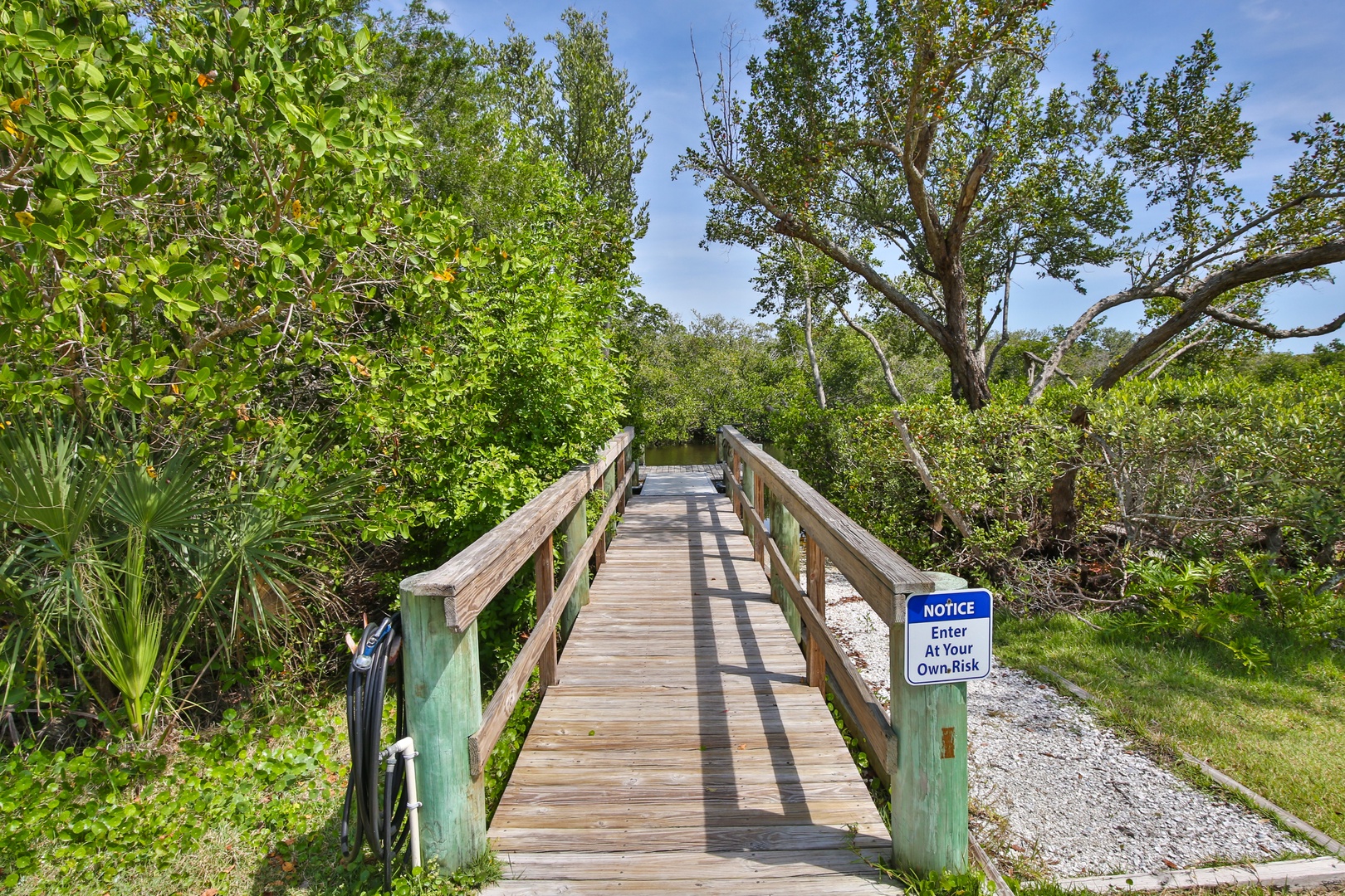A wooden footbridge leads through lush tropical vegetation toward the property, surrounded by mature trees and native greenery. The sunny day highlights the natural setting.